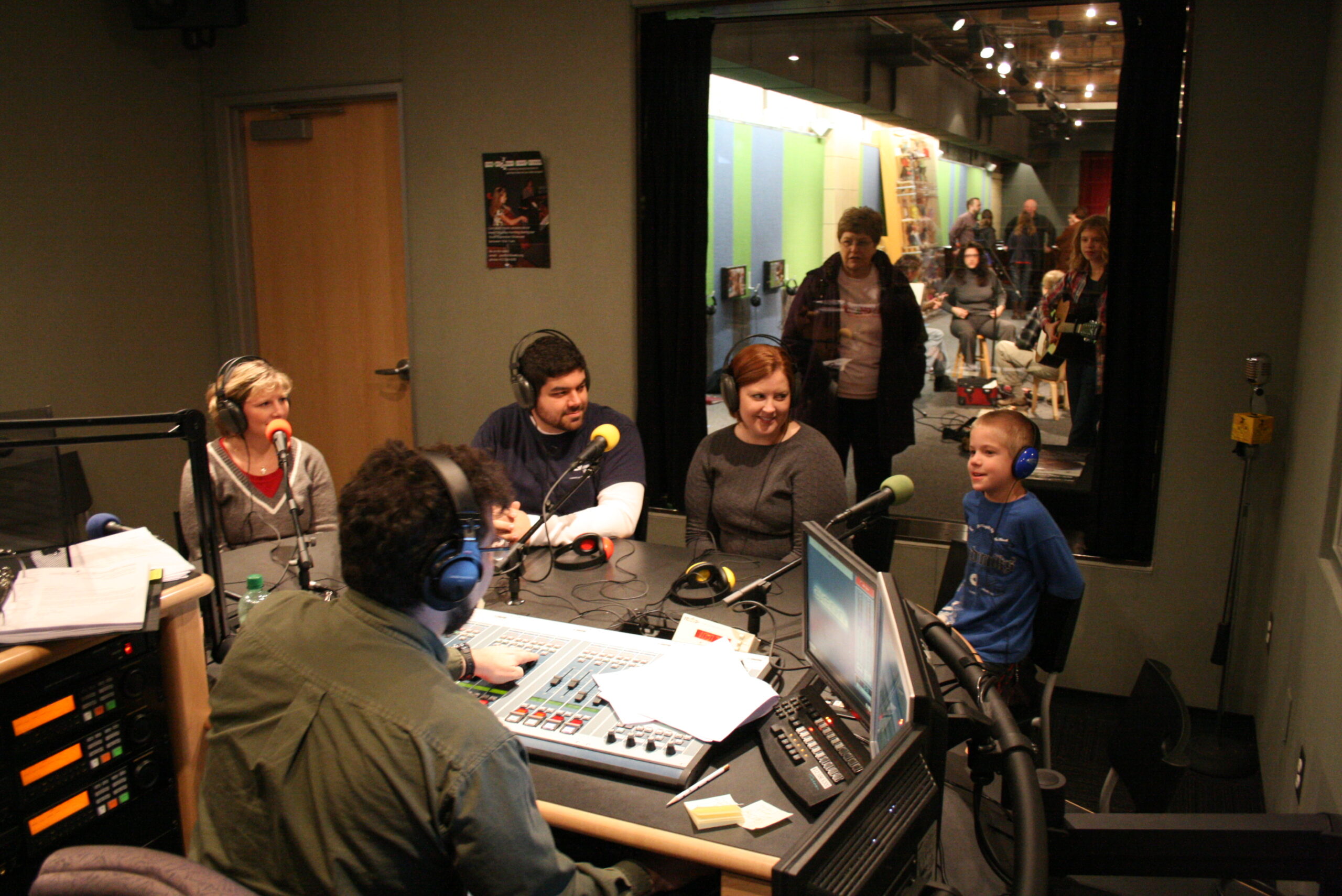 Three adults and a youth wearing headphones speaking into microphones being interviewed by an adult wearing headphones sitting behind an audio mixer in a radio studio