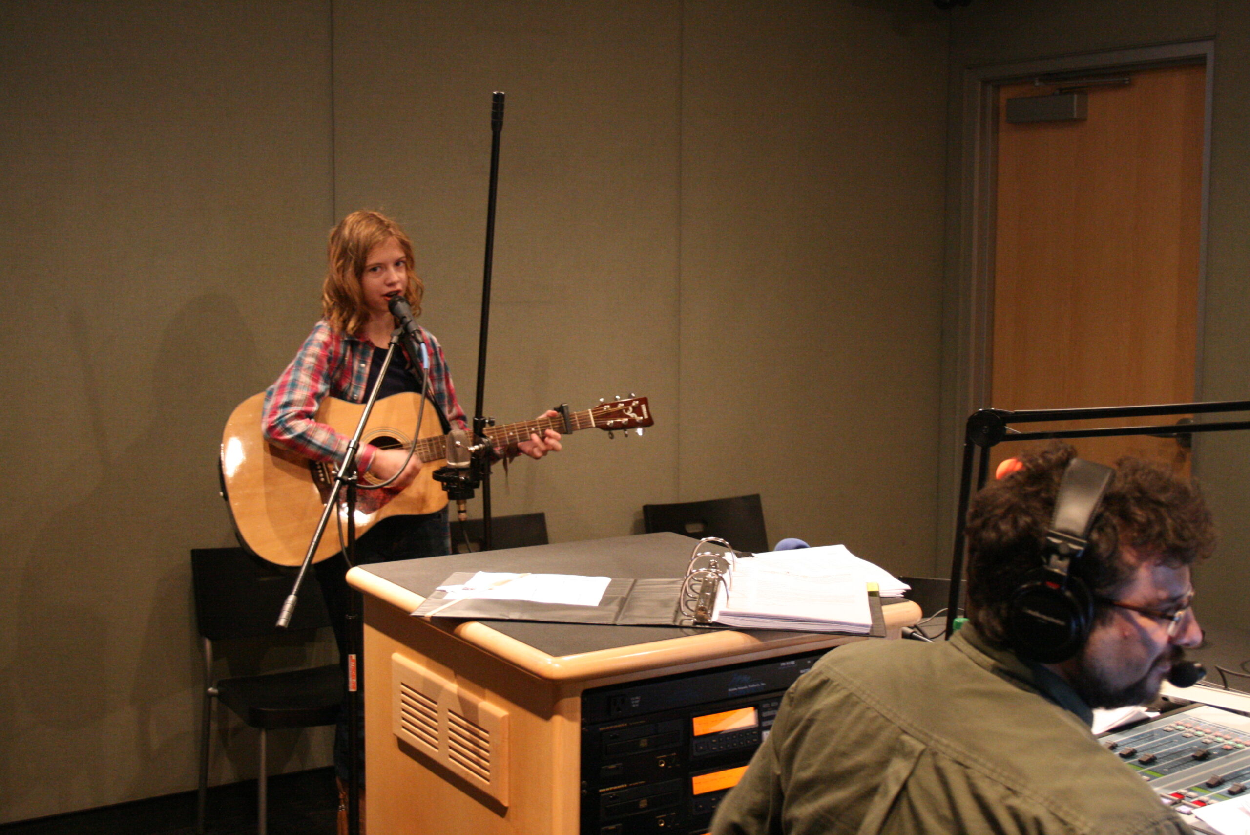 A teen playing an acoustic guitar and singing into a microphone as an adult wearing headphones listens in a radio studio