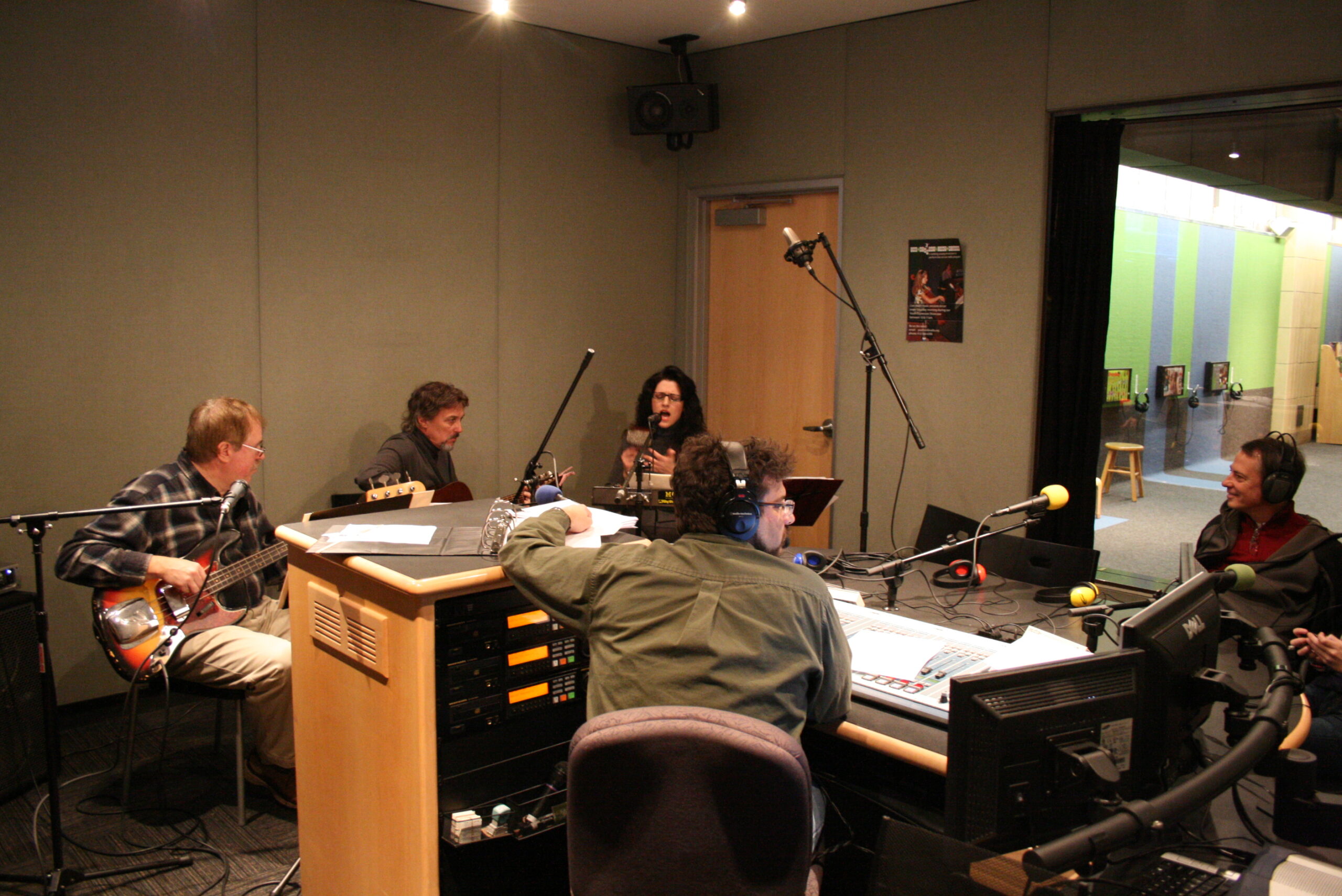 An adult playing an electric bass, an adult holding an acoustic guitar, and three adults sitting behind microphones as an adult wearing headphones sitting behind an audio mixer listens in a radio studio