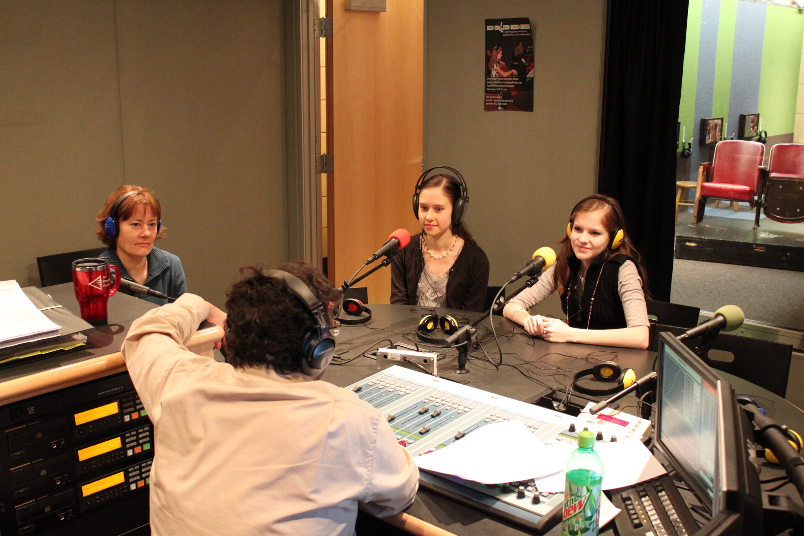 Two teens and an adult wearing headphones sitting behind microphones being interviewed by an adult wearing headphones sitting behind an audio mixer in a radio studio