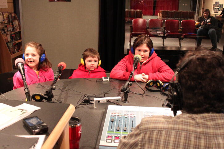 Three youth wearing headphones speaking into microphones being interviewed by an adult wearing headphones in a radio studio