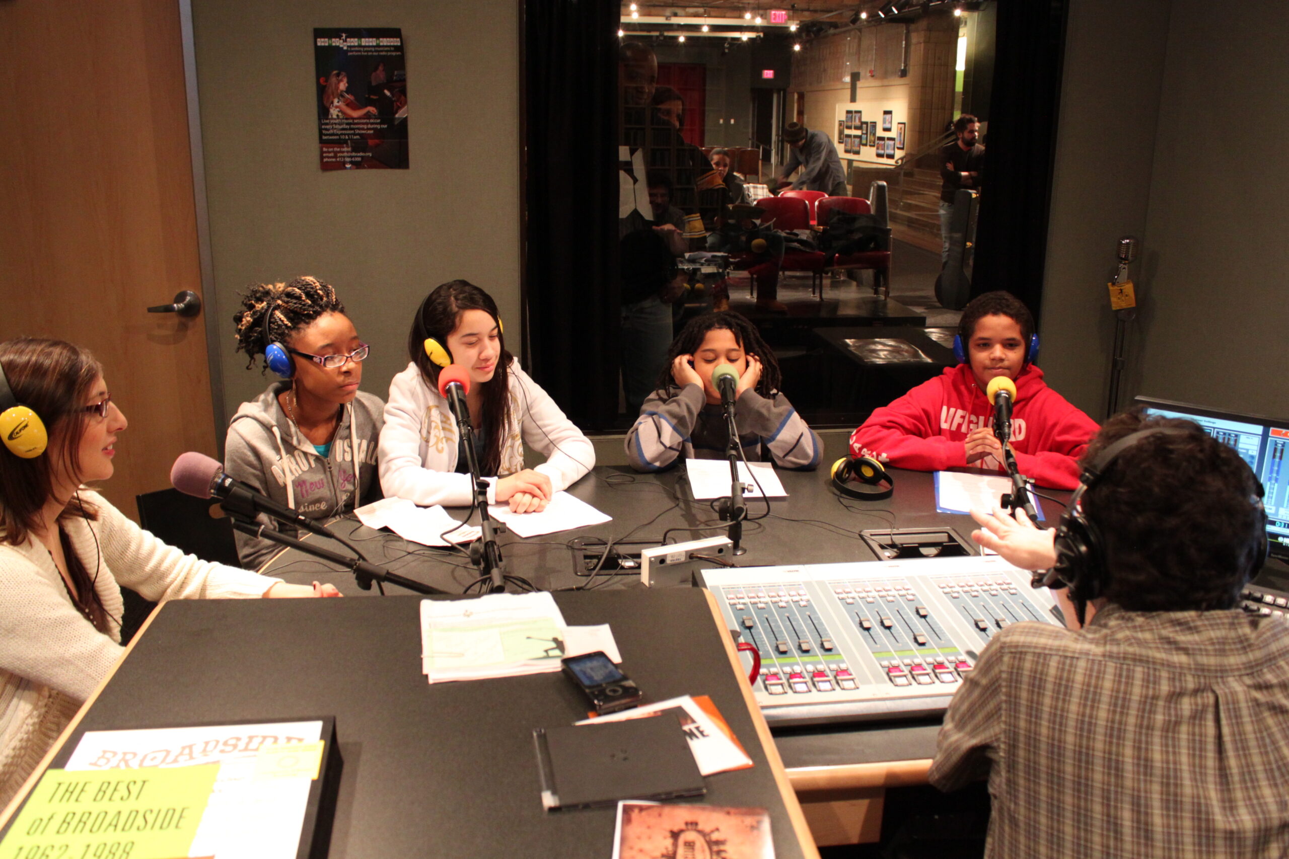 Two adults and three youth wearing headphones sitting behind microphones being interviewed by an adult wearing headphones sitting behind an audio mixer in a radio studio