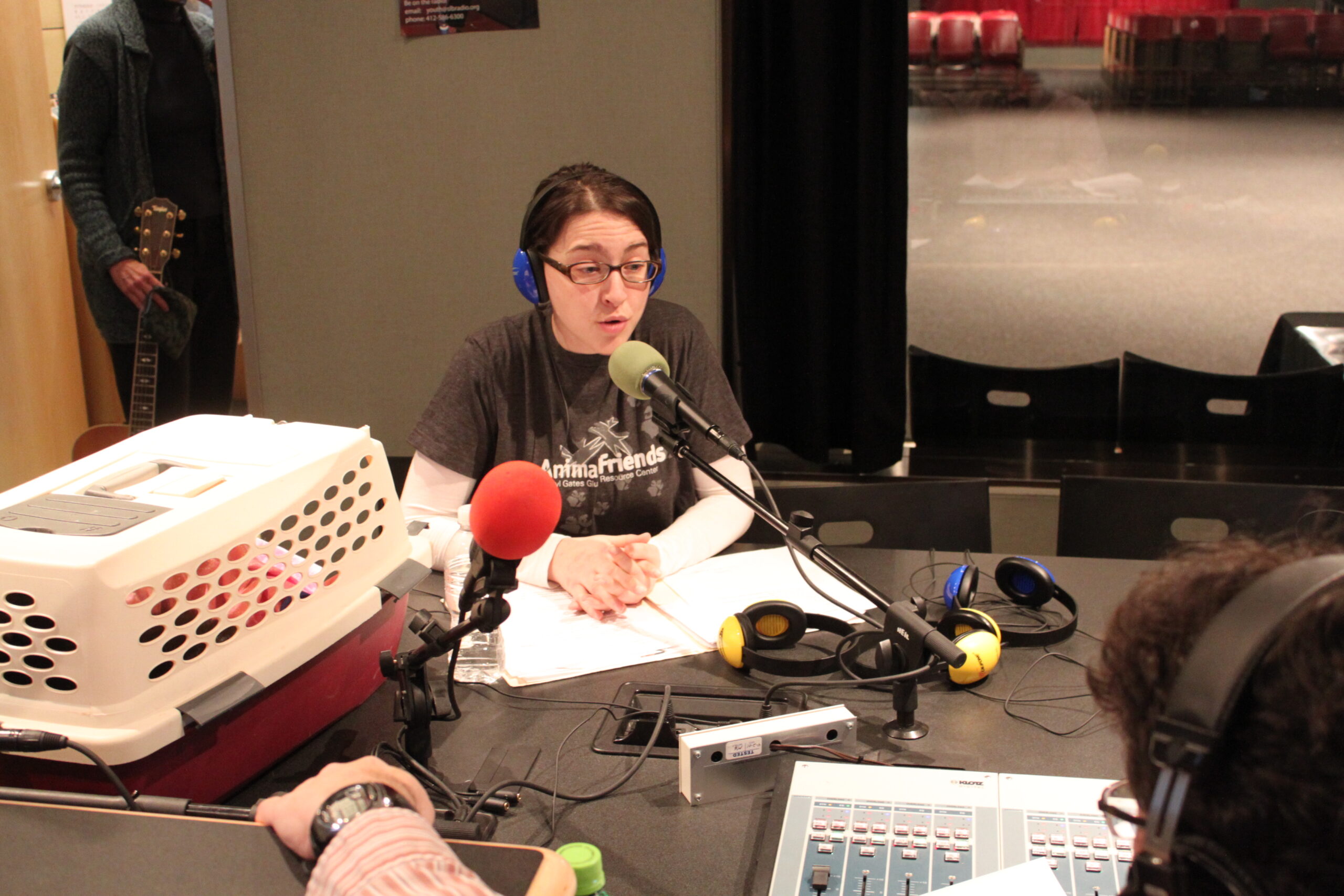 An adult wearing headphones sitting next to a pet carrier and speaking into a microphone in a radio studio