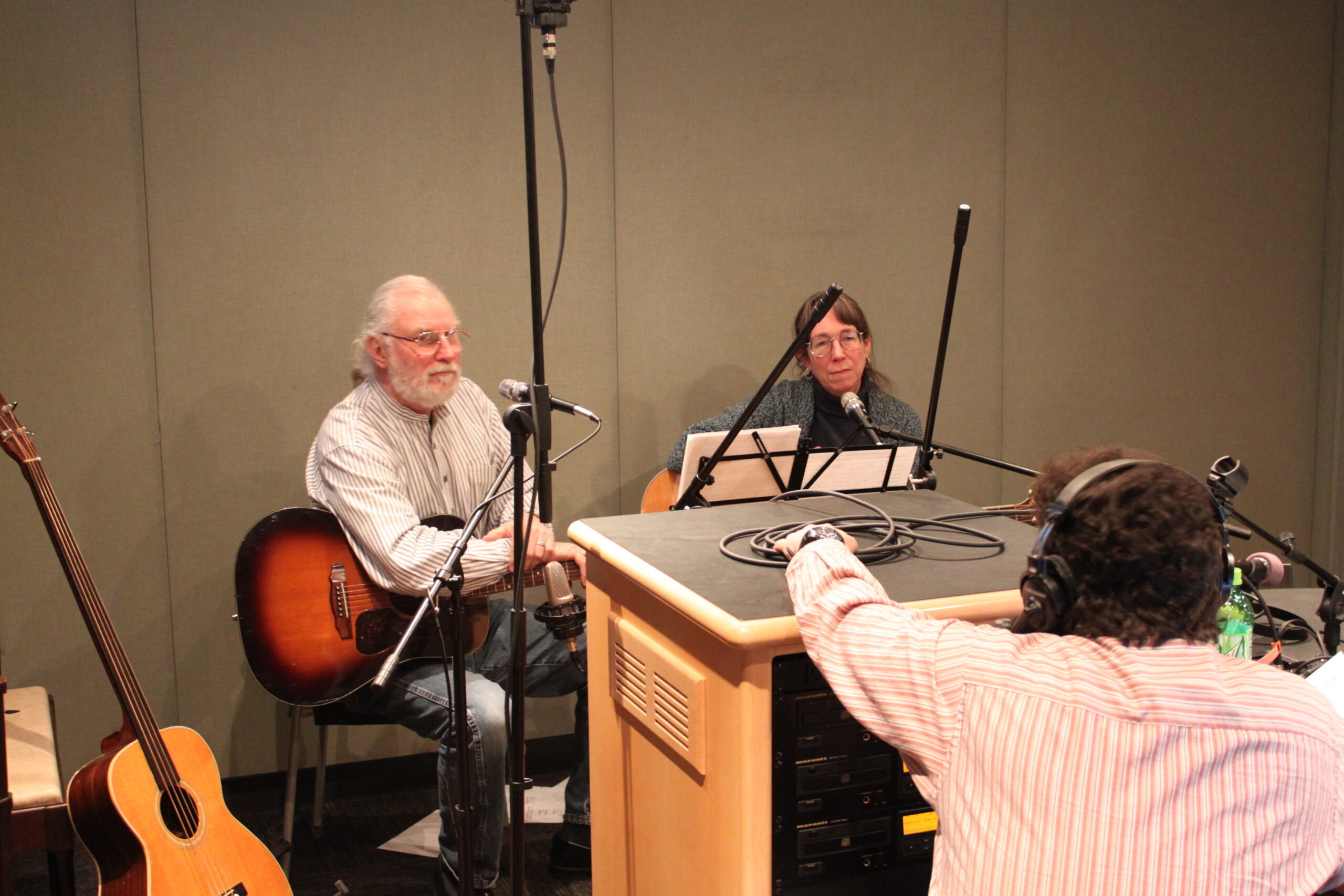 Two adults holding acoustic guitars sitting behind microphones being interviewed by an adult wearing headphones in a radio studio