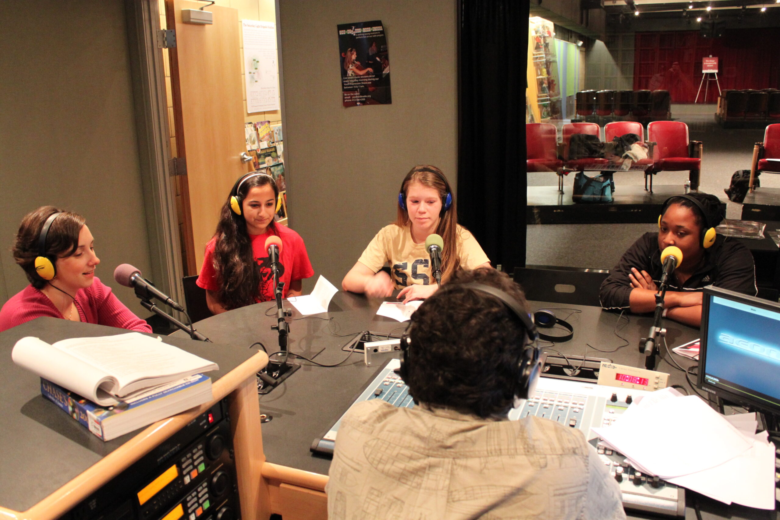 Three teens and an adult wearing headphones speaking into microphones being interviewed by an adult wearing headphones in a radio studio