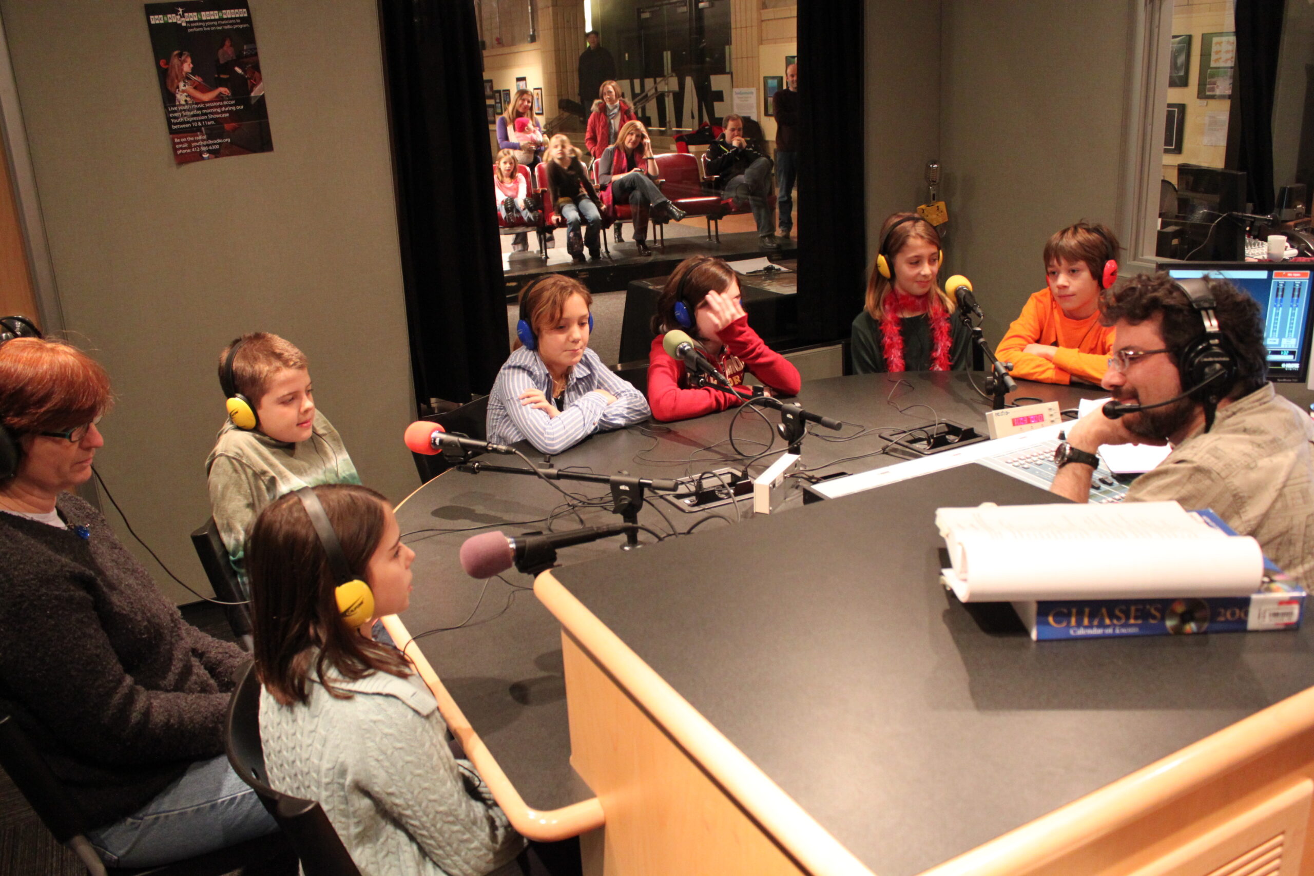 Six youth and one adult wearing headphones sitting behind microphones being interviewed by an adult wearing headphones sitting behind a microphone and an audio mixer in a radio studio
