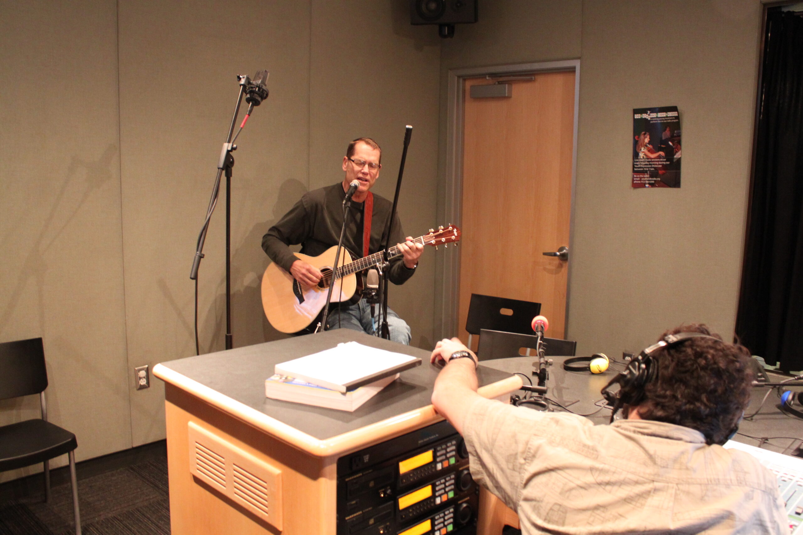 An adult playing an acoustic guitar and singing into a microphone as an adult wearing headphones listens in a radio studio