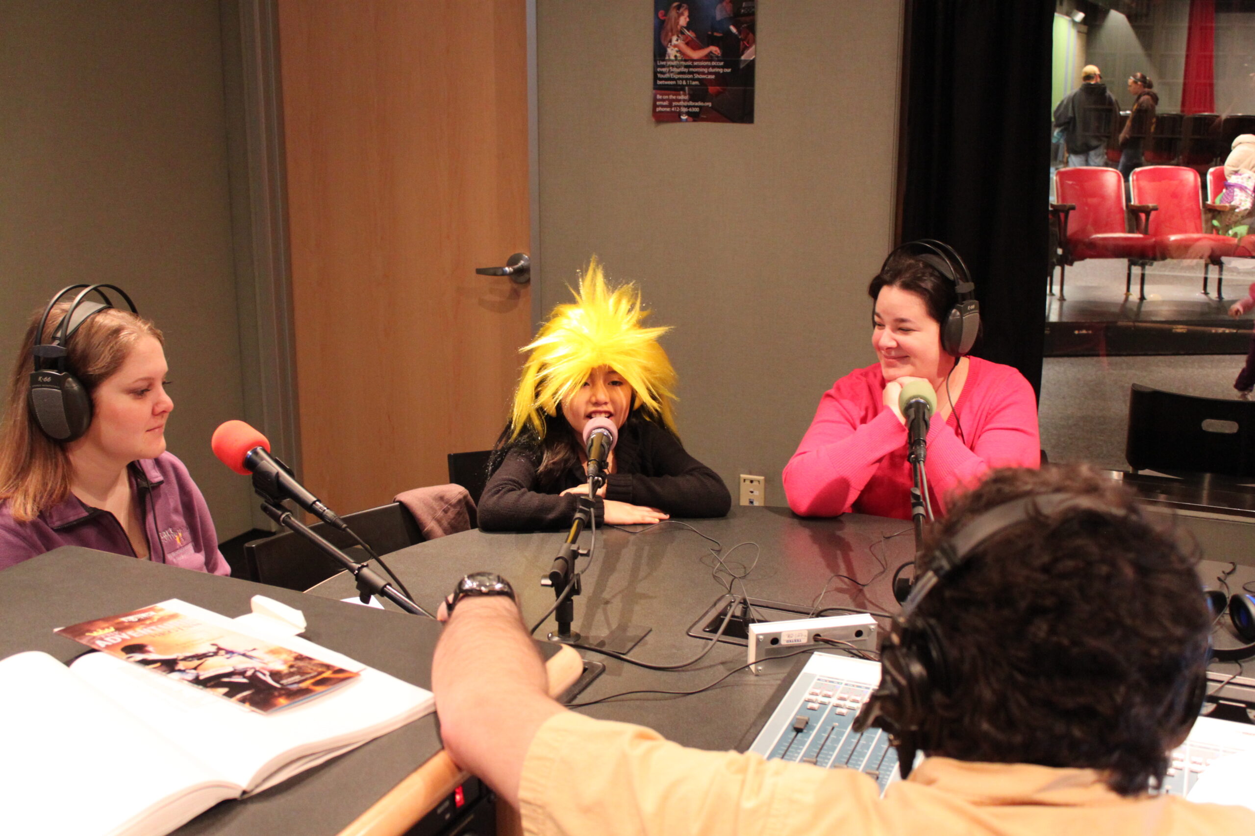 Two adults and a youth wearing headphones speaking into microphones being interviewed by an adult wearing headphones in a radio studio