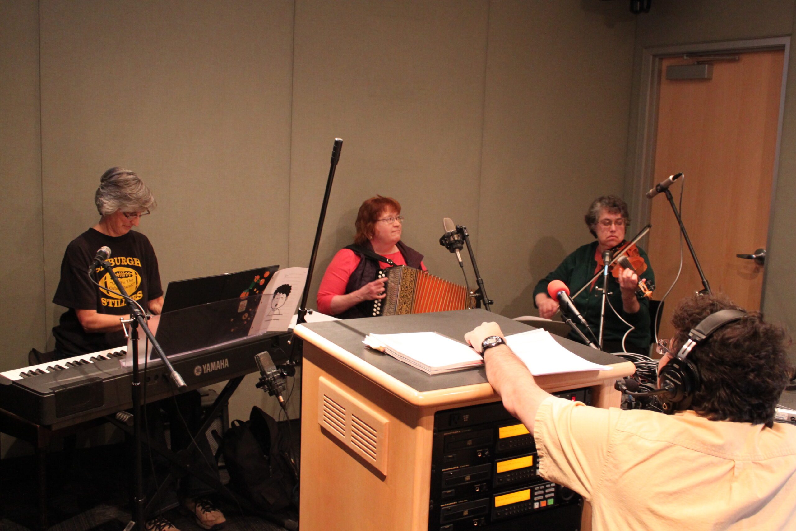 An adult playing a piano keyboard, an adult playing an accordion, and an adult playing a violin as an adult wearing headphones listens in a radio studio