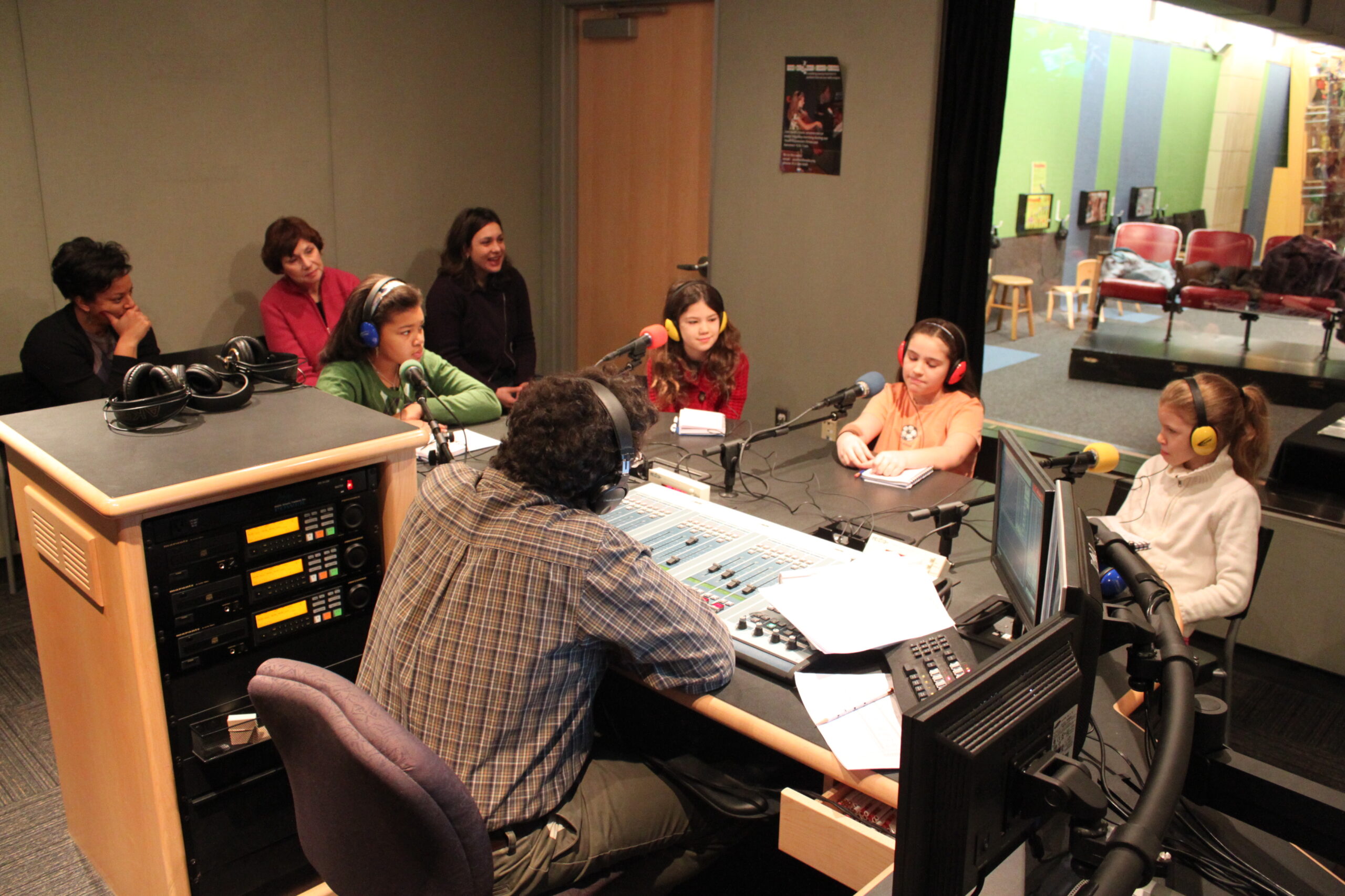 Seven teens, four wearing headphones, sitting behind microphones being interviewed by an adult wearing headphones sitting behind an audio mixer in a radio studio