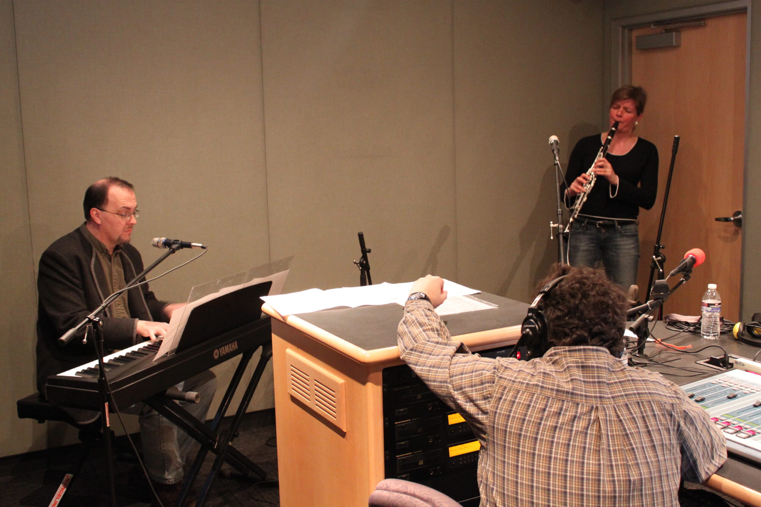 An adult playing a piano keyboard and an adult playing clarinet as an adult wearing headphones listens in a radio studio