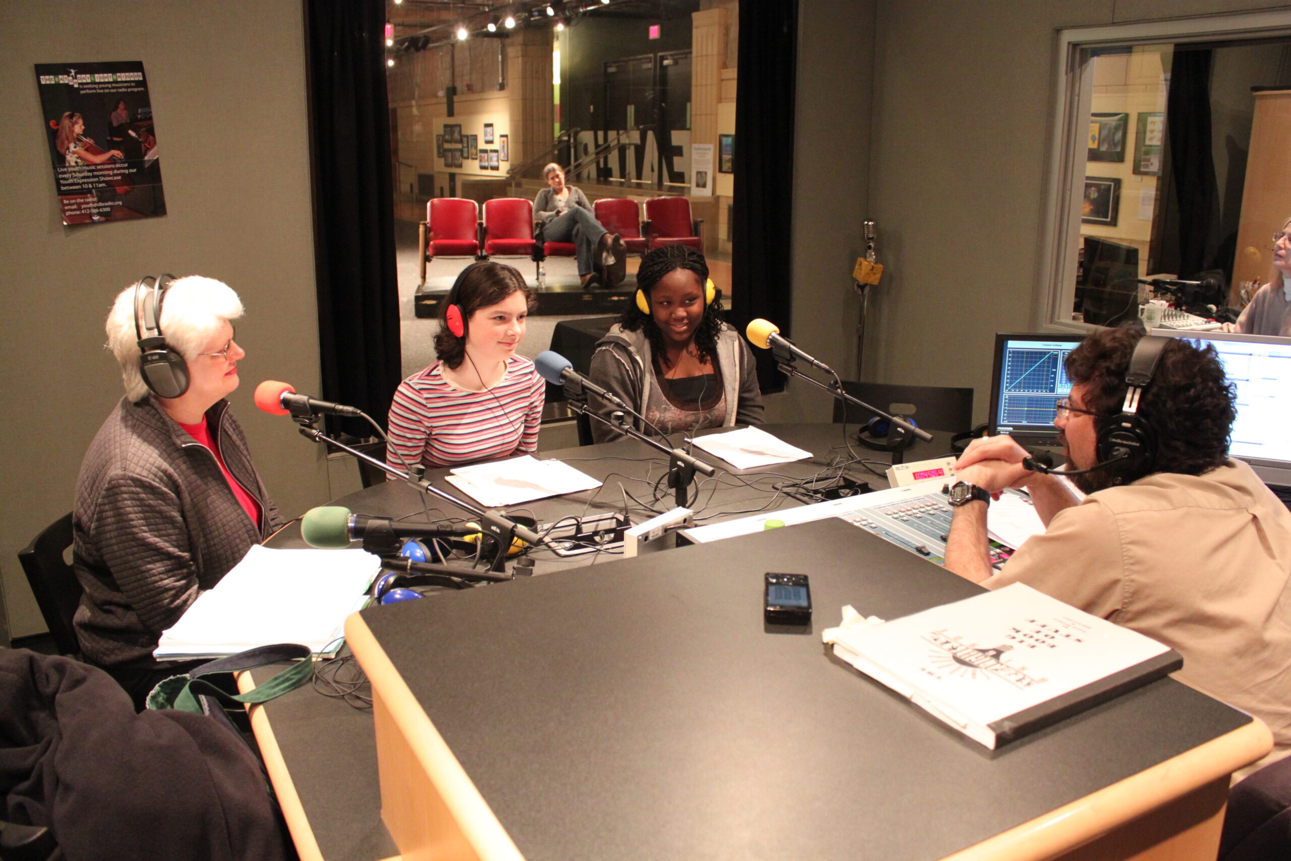 Two teens and an adult wearing headphones speaking into microphones being interviewed by an adult wearing headphones sitting behind an audio mixer