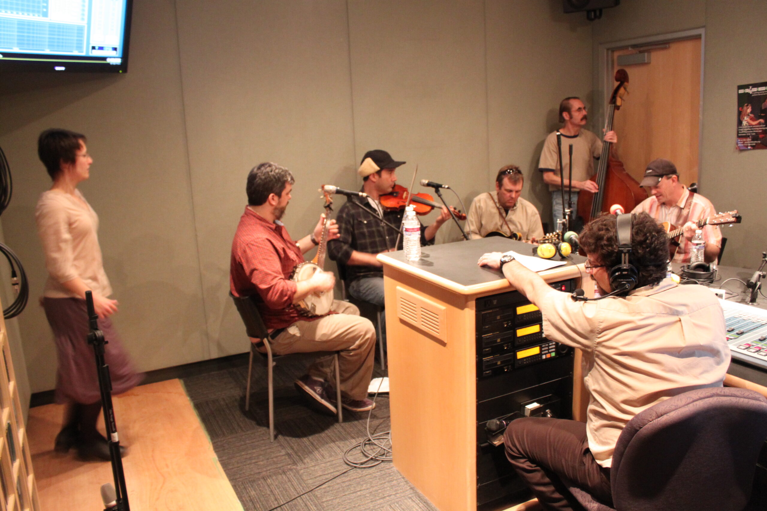 An adult playing banjo, an adult playing violin, an adult playing a string instrument, an adult playing upright bass, and an adult playing acoustic guitar as two adults watch in a radio studio