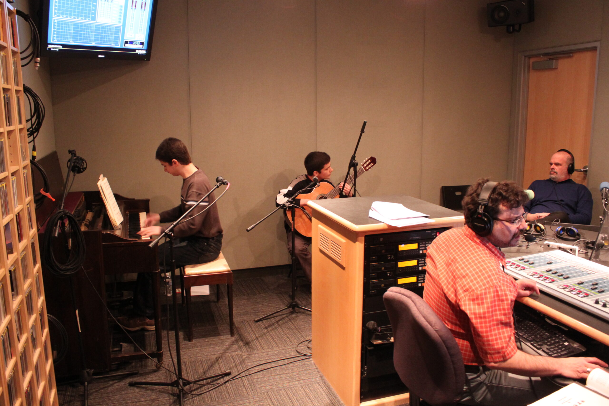 A teen playing a piano keyboard and a teen playing an acoustic guitar as two adults wearing headphones listen in a radio studio
