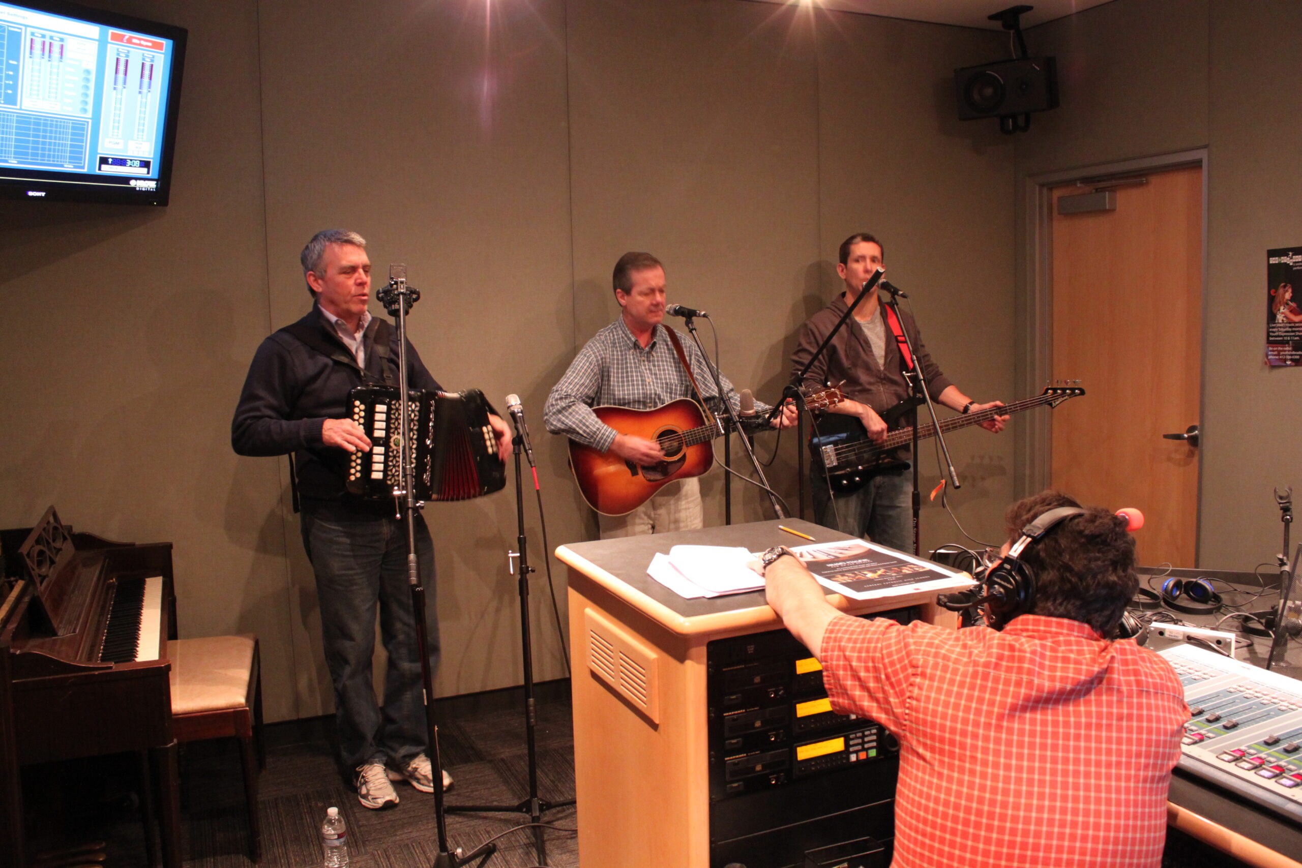 An adult playing accordion, an adult playing acoustic guitar, and an adult playing electric bass as an adult wearing headphones listening in a radio studio