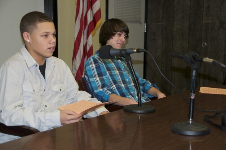 Two teens sitting behind a microphone on a brown table