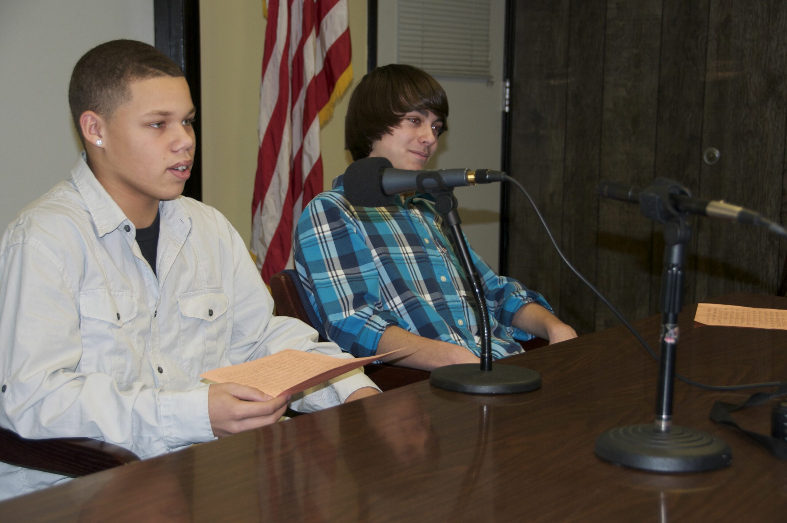 Two teens sitting behind a microphone on a brown table