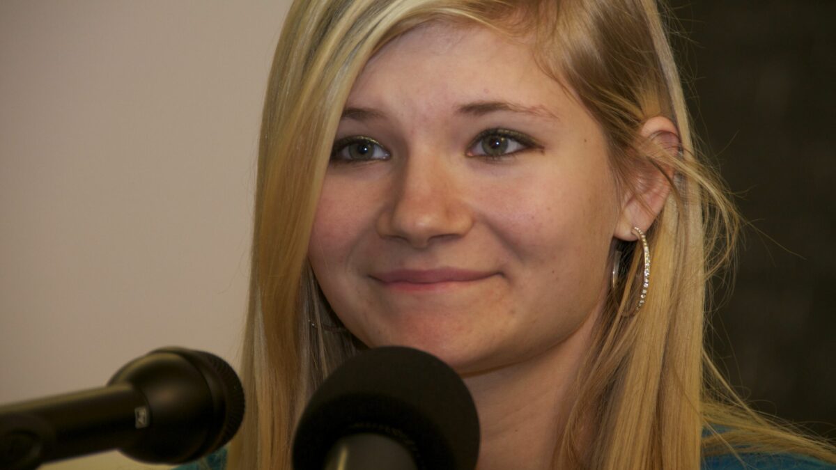 A teen with long blonde hair sitting behind two microphones