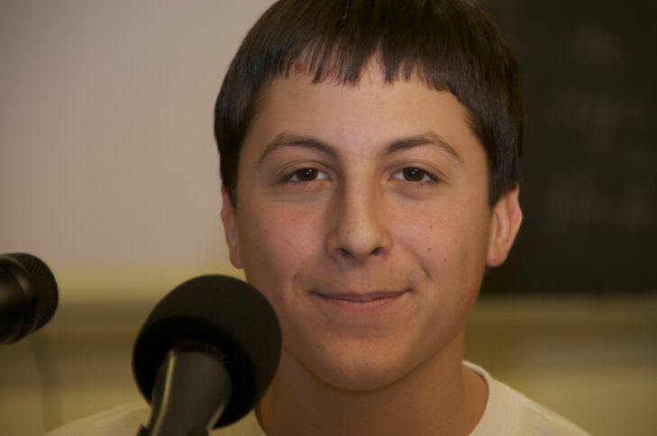 A teenager with short brown hair smiling behind a microphone