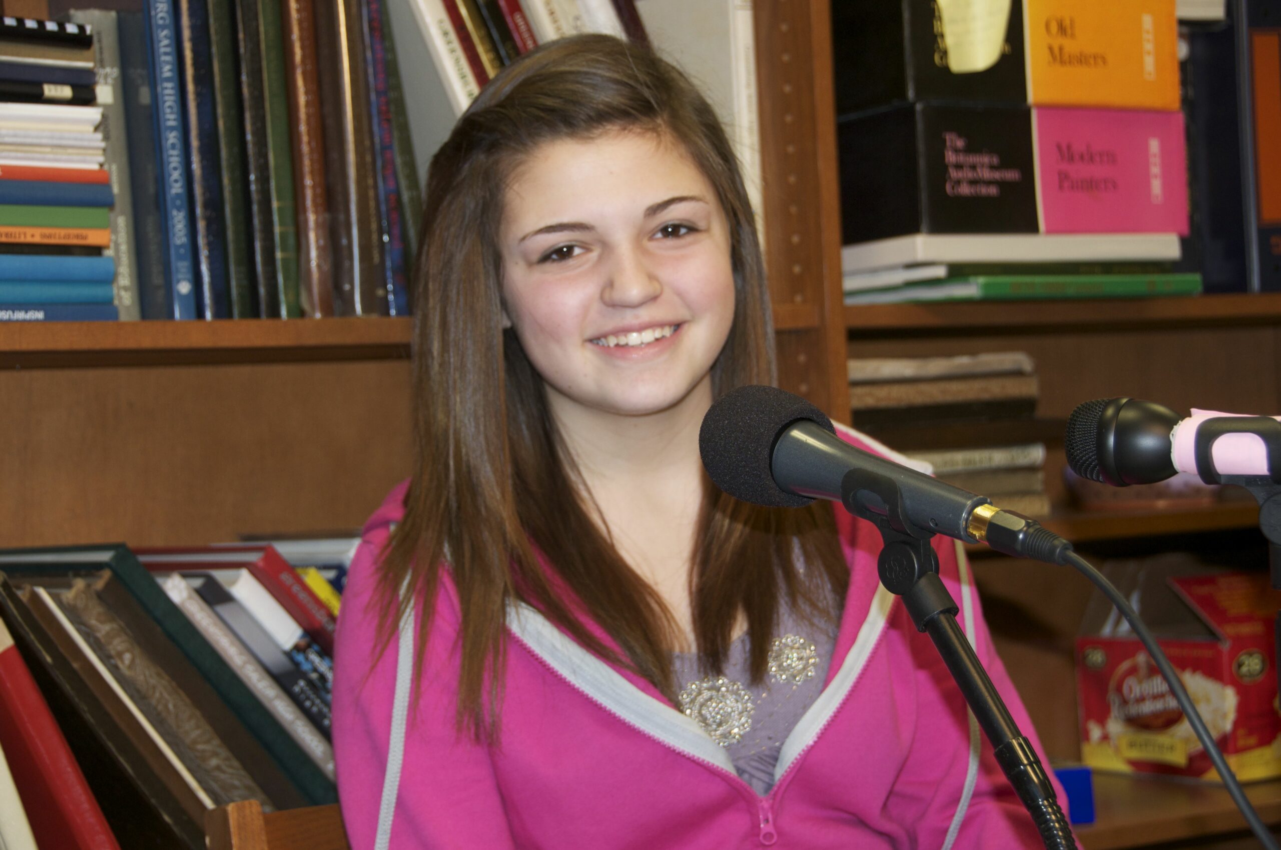 A teen smiling while sitting behind a microphone in front of a bookshelf