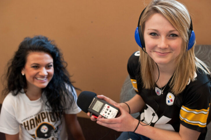 A teen wearing headphones interviewing another teen with a portable recorder