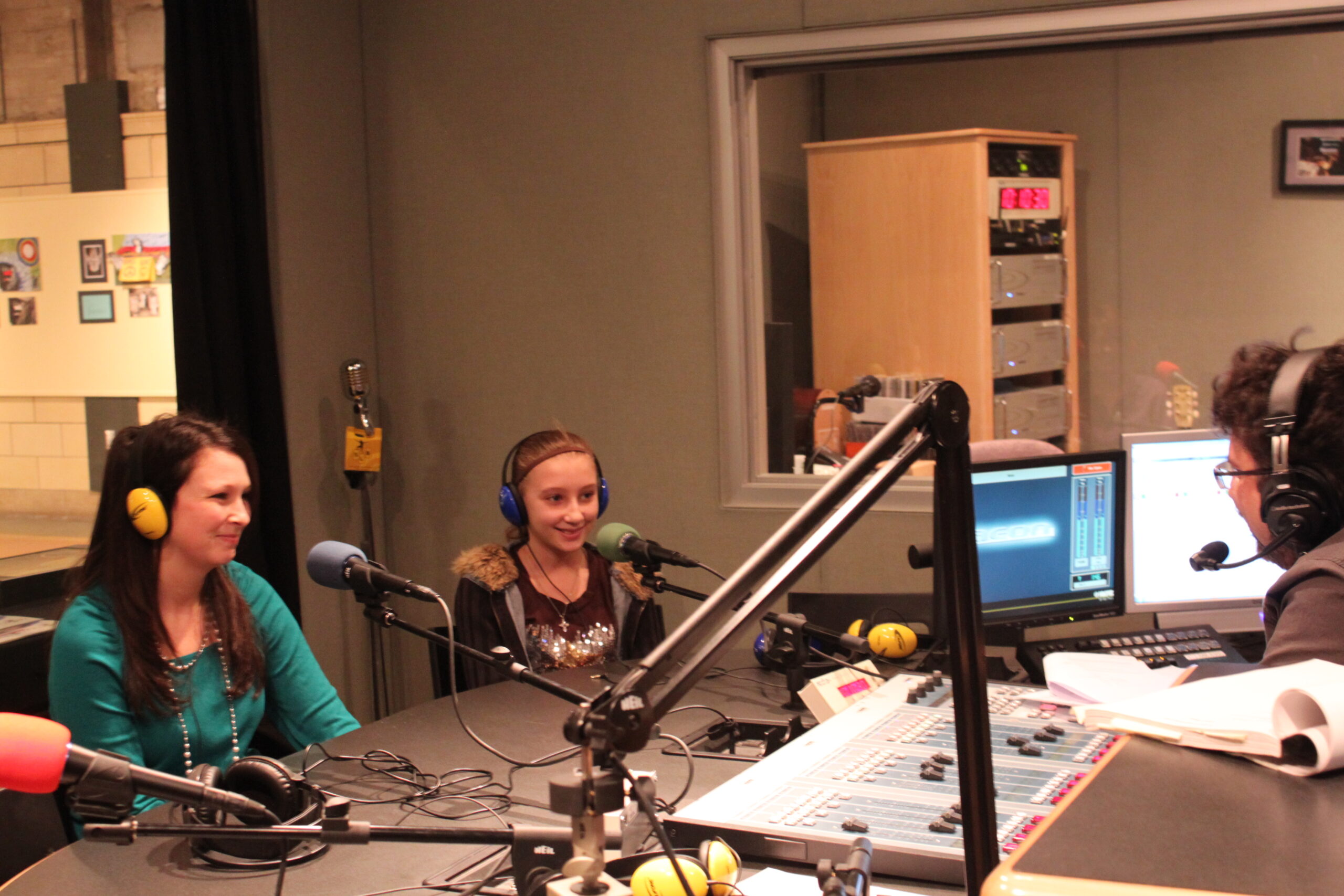An adult and a youth wearing headphones sitting behind microphones being interviewed by an adult wearing headphones sitting behind an audio mixer in a radio studio