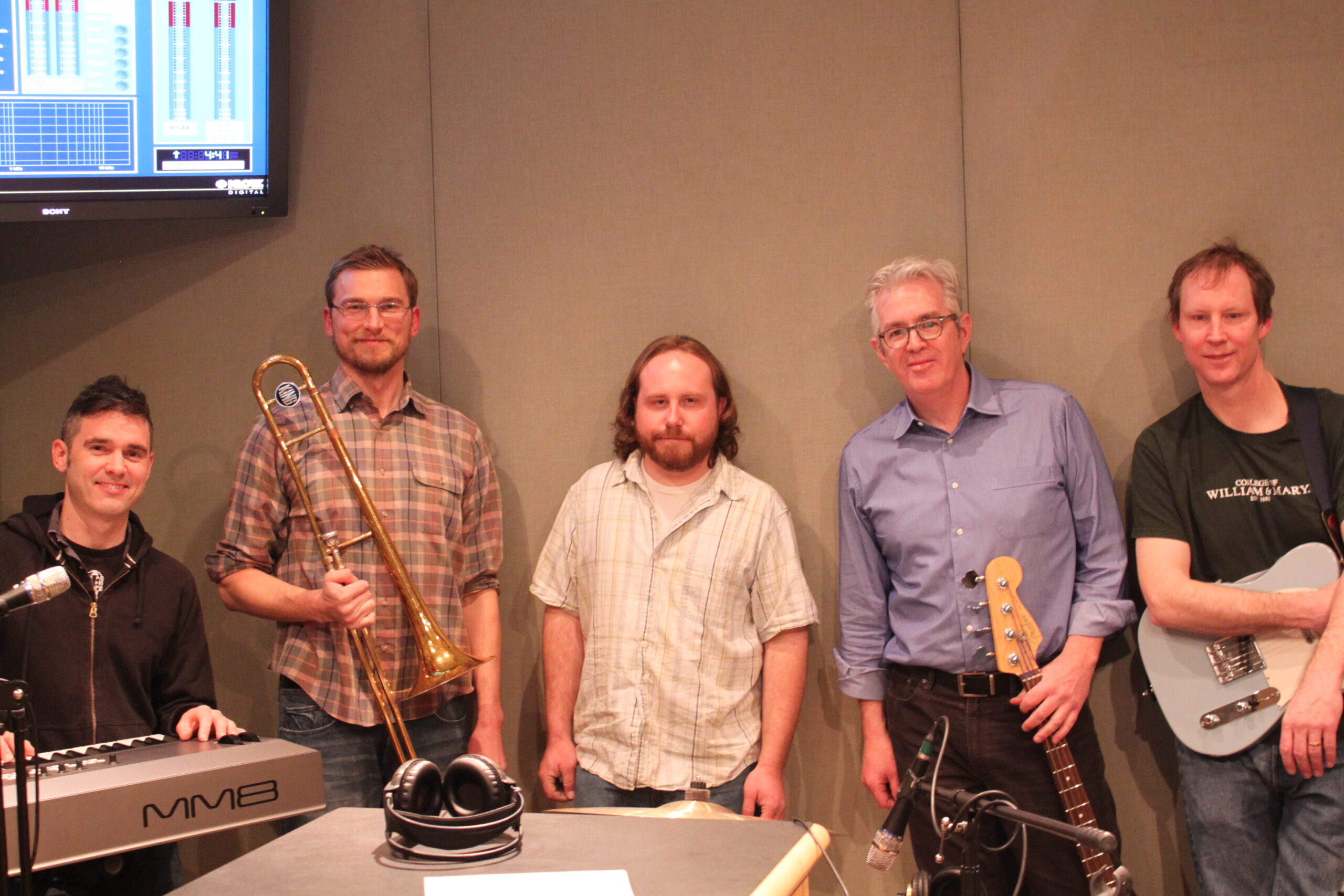 Five adults smiling and posing while holding various instruments in a radio studio