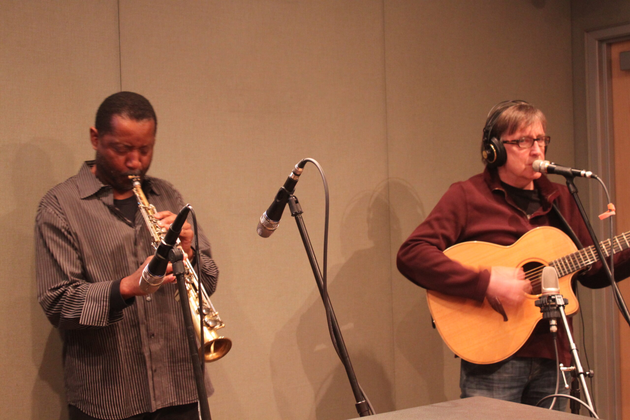 An adult playing a soprano saxophone and an adult playing an acoustic guitar in a radio studio