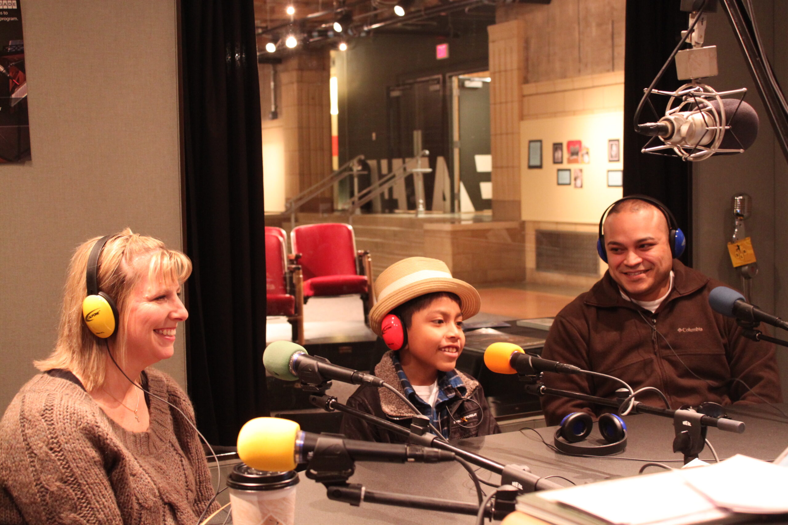Two adults and a youth smiling while wearing headphones and sitting behind microphones in a radio studio