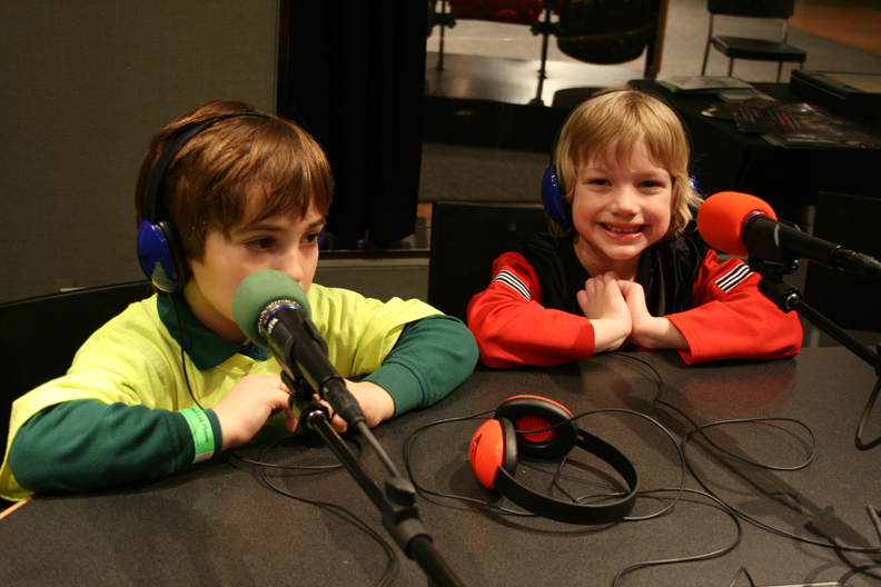 Two youth wearing headphones sitting behind microphones in a radio studio