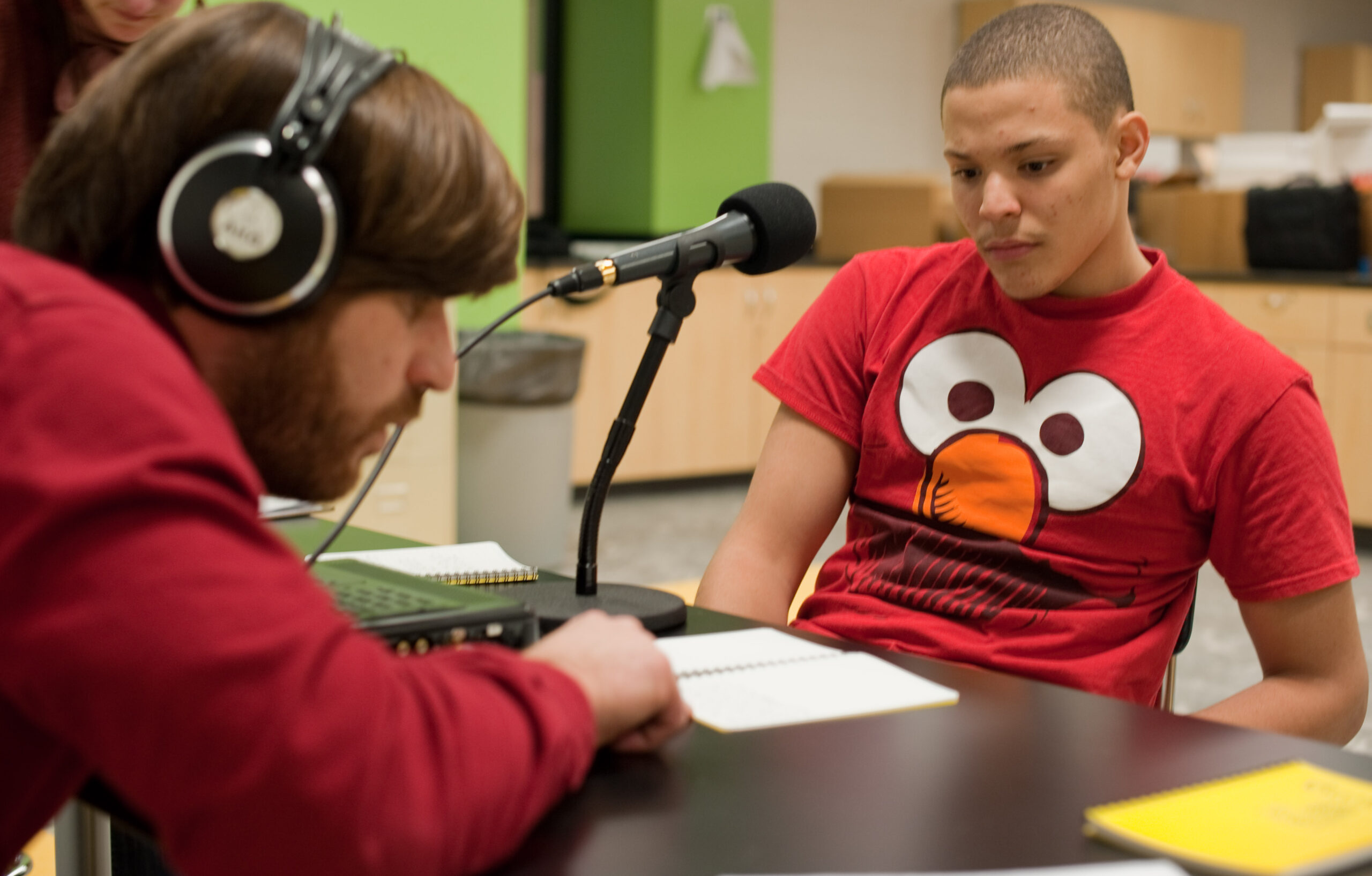 An adult wearing headphones interviewing a teen sitting behind a microphone