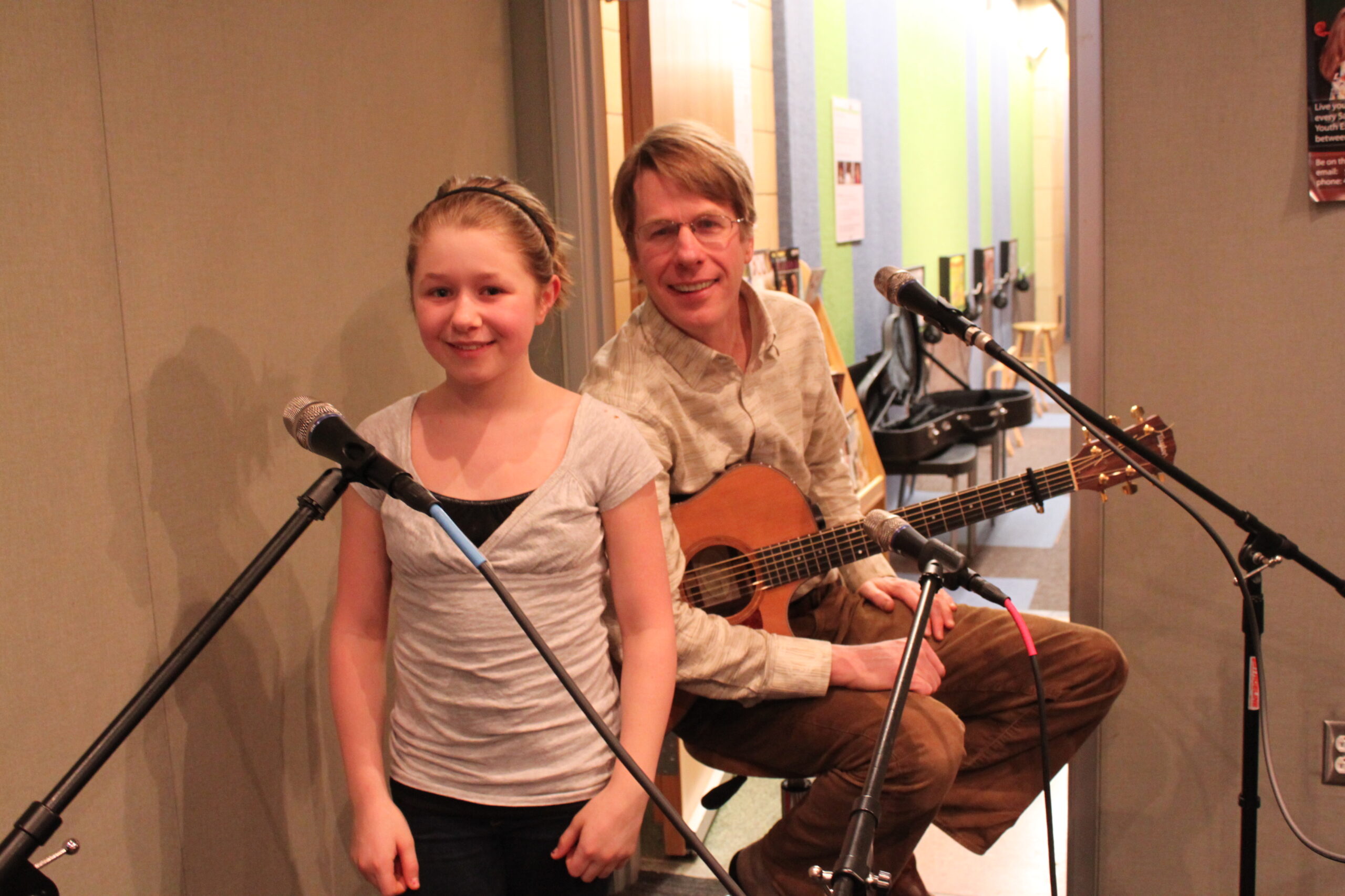 An adult holding an acoustic guitar and a youth, both behind microphones in a radio studio