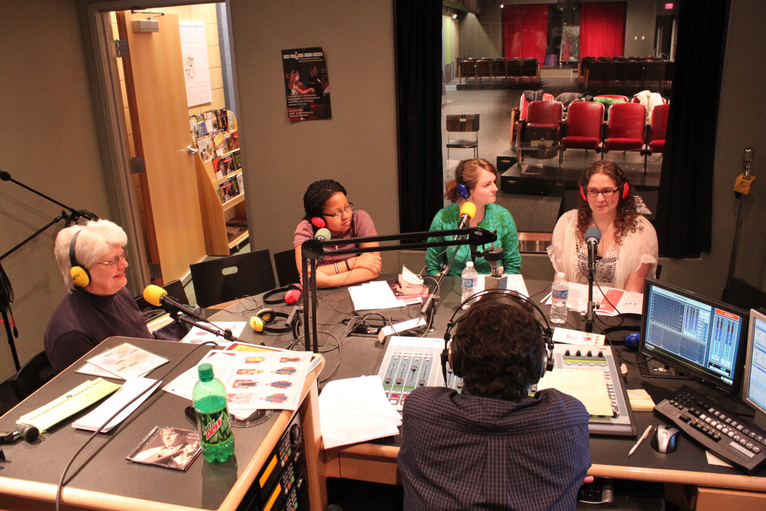 An adult and three teens wearing headphones speaking into microphones being interviewed by an adult wearing headphones sitting behind an audio mixer in a radio studio