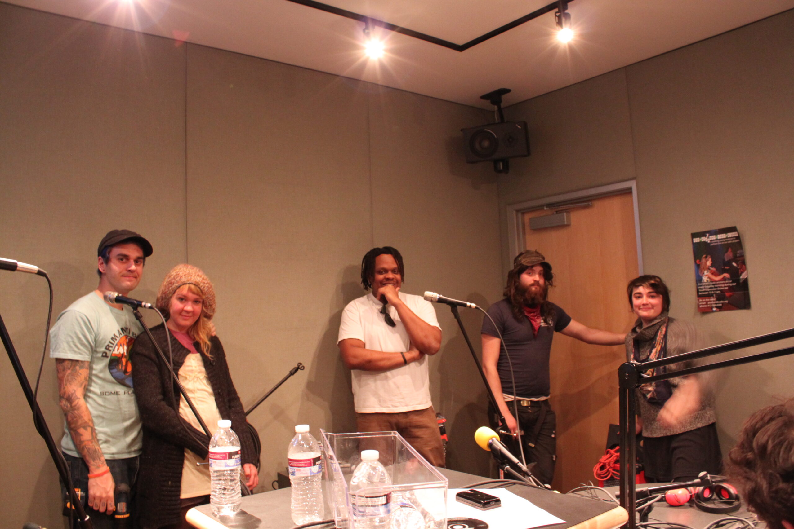 Five adults standing behind microphones in a radio studio