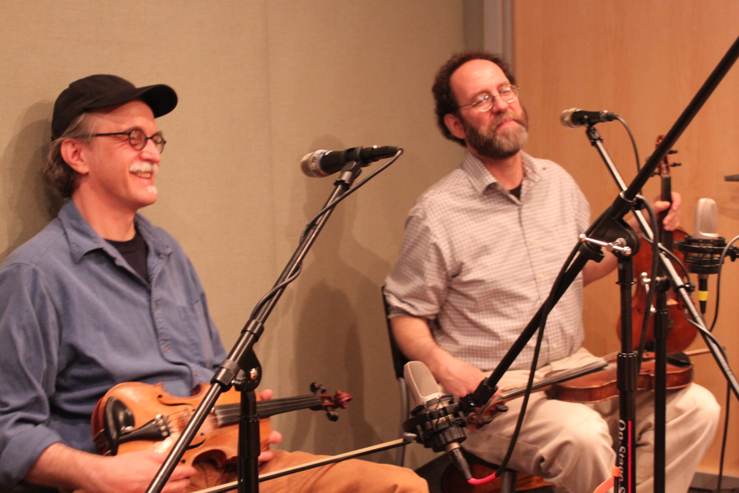 Two adults holding violins and sitting behind microphones in a radio studio