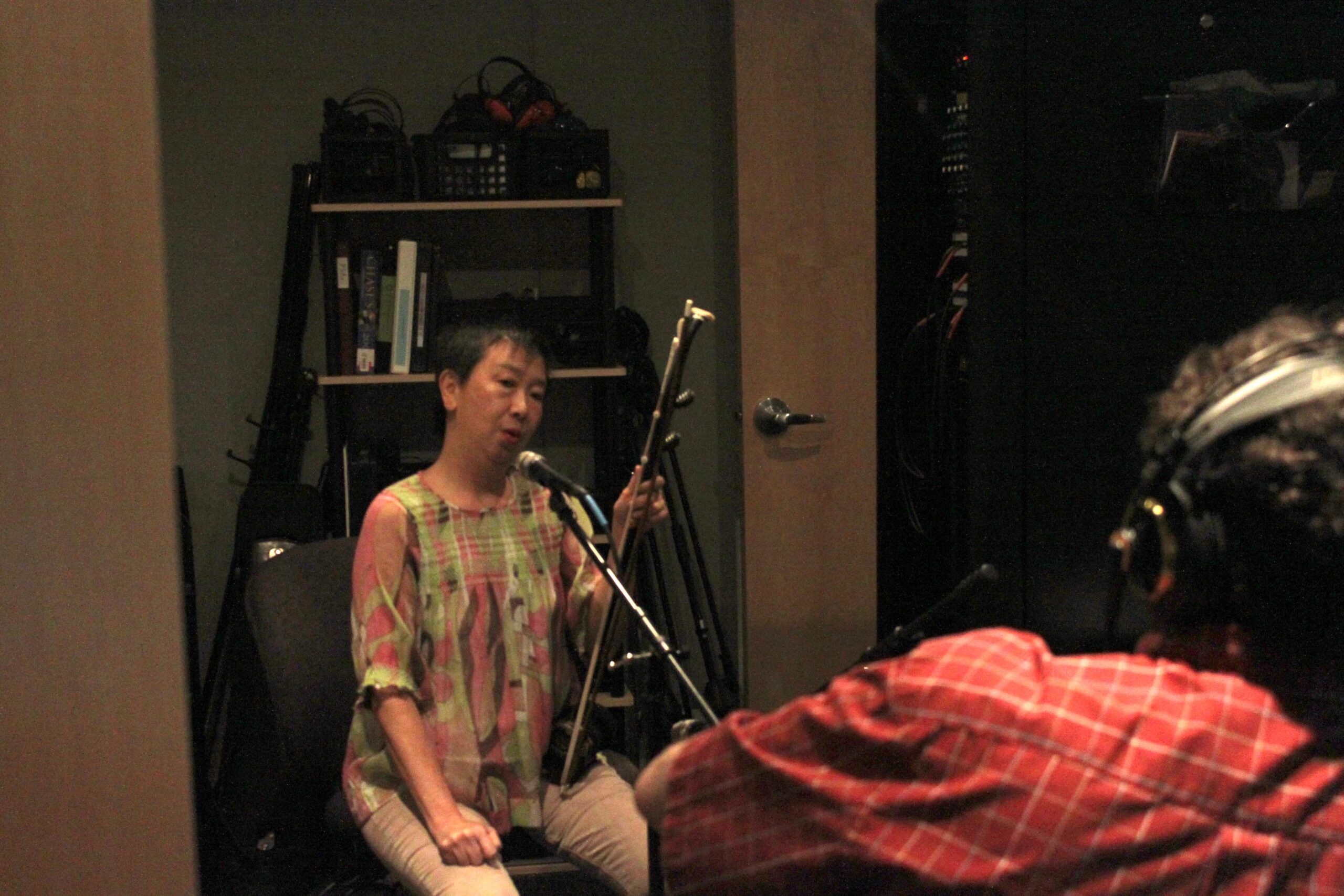 An adult holding an ethnic instrument speaking into a microphone being interviewed by an adult wearing headphones in a radio studio