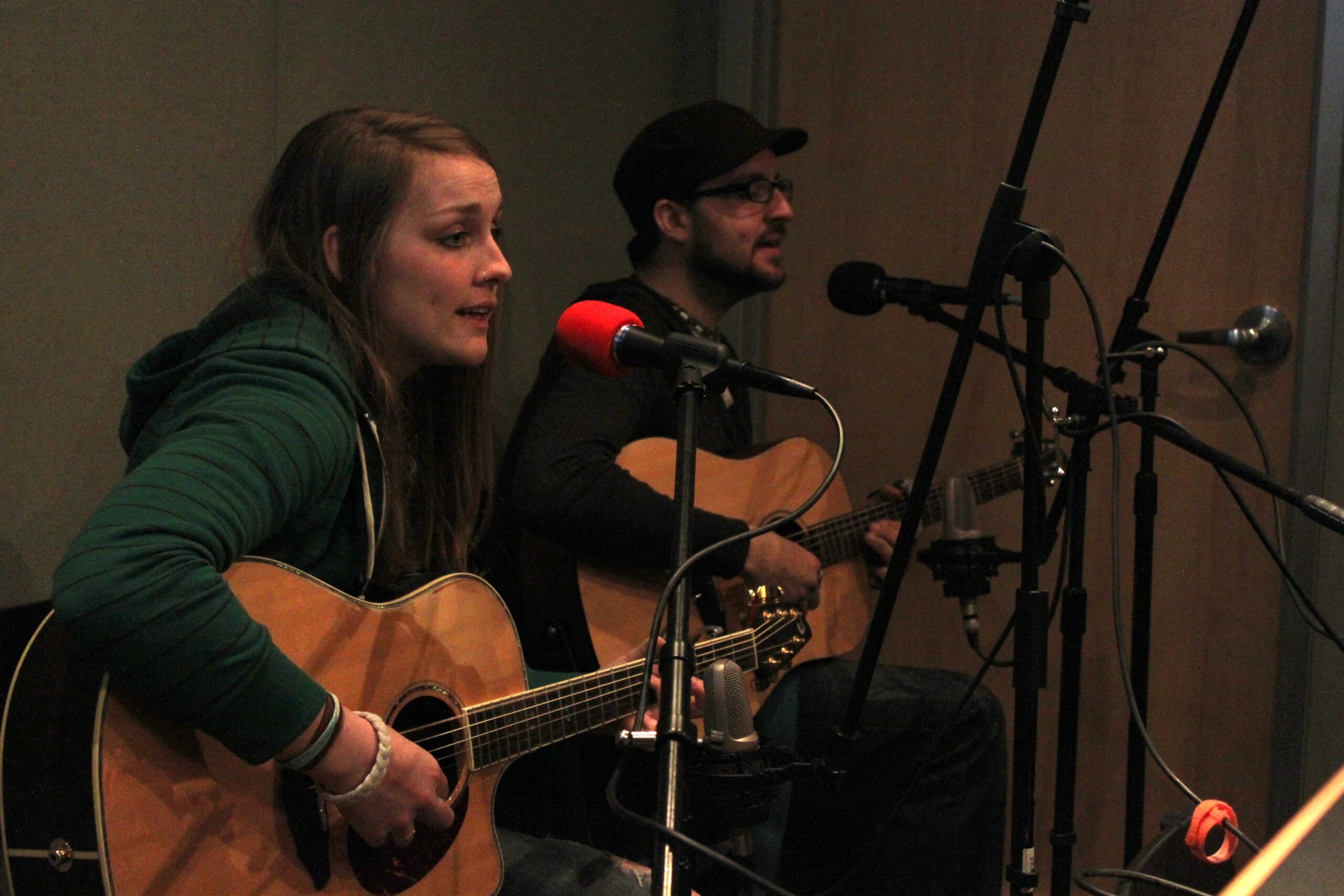 Two adults playing acoustic guitars and singing into microphones in a radio studio