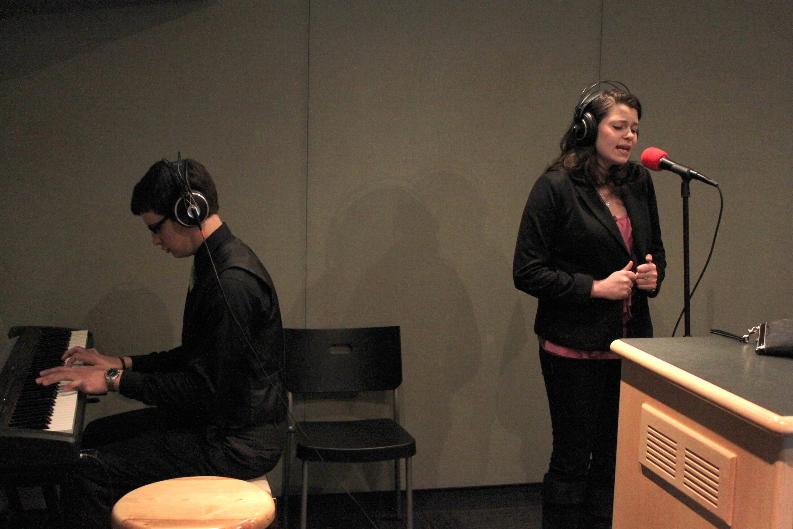 An adult playing a piano keyboard and a teen singing into a microphone in a radio studio