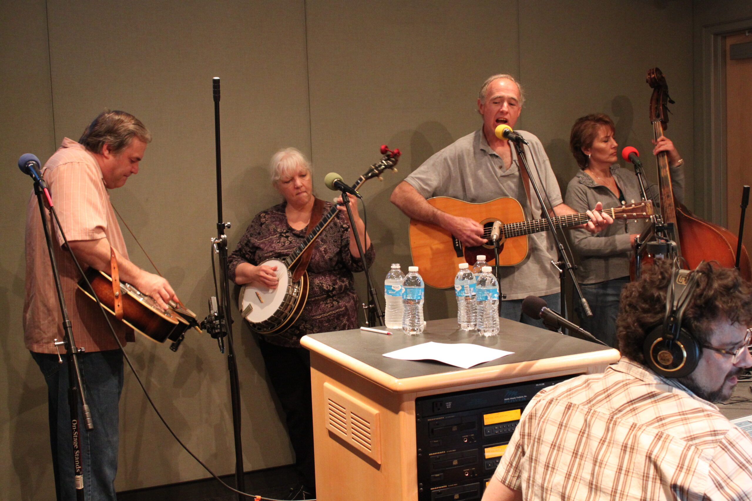 An adult playing a dobro, an adult playing a banjo, an adult playing an acoustic guitar, and an adult playing an upright bass as an adult wearing headphones listens in a radio studio