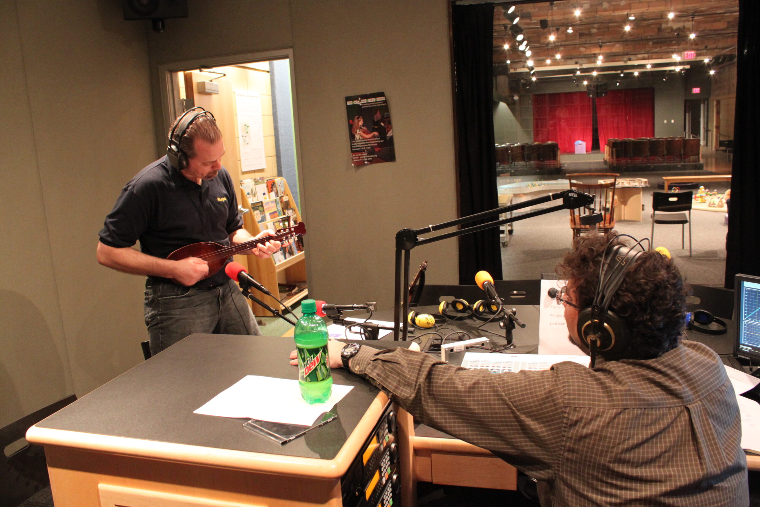An adult wearing headphones playing a small ethnic string instrument as another adult wearing headphones listens in a radio studio