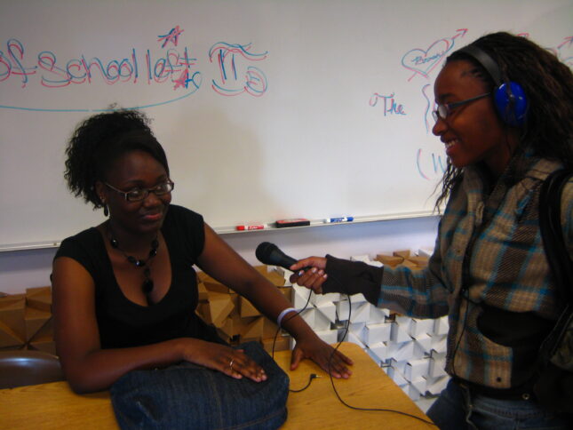 A teen wearing headphones interviewing another teen speaking into a microphone in a classroom