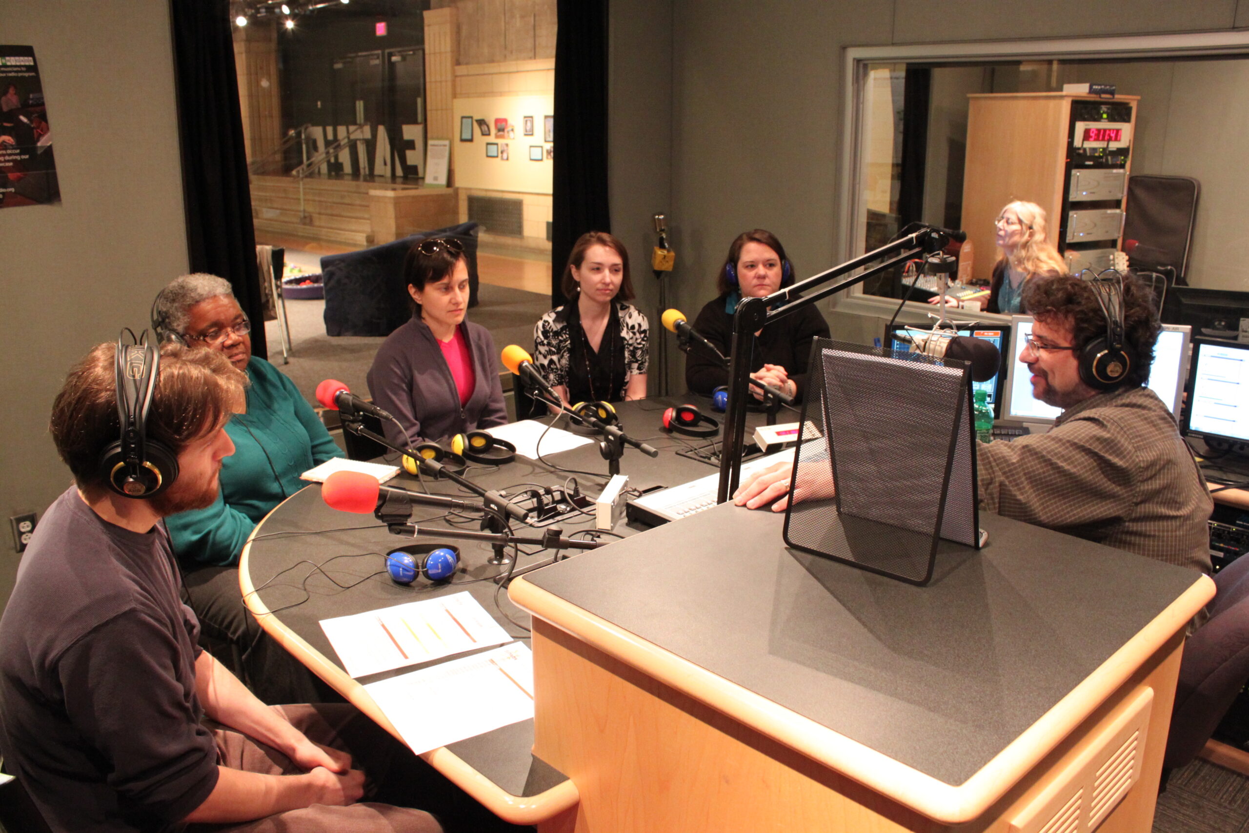Five adults wearing headphones sitting behind microphones being interviewed by an adult wearing headphones sitting behind a microphone in a radio studio
