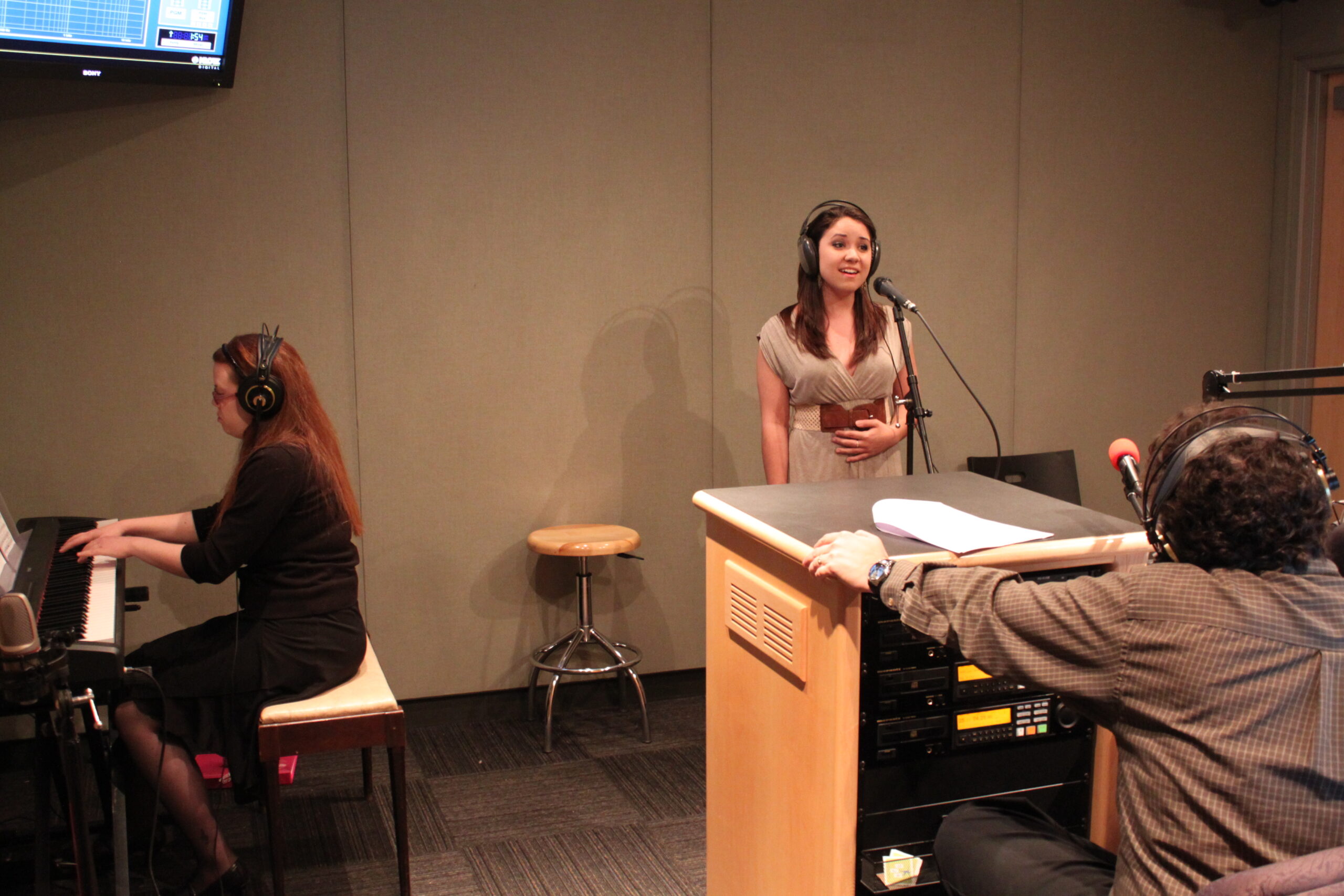 An adult playing a piano keyboard and a teen singing into a microphone as an adult wearing headphones listens in a radio studio
