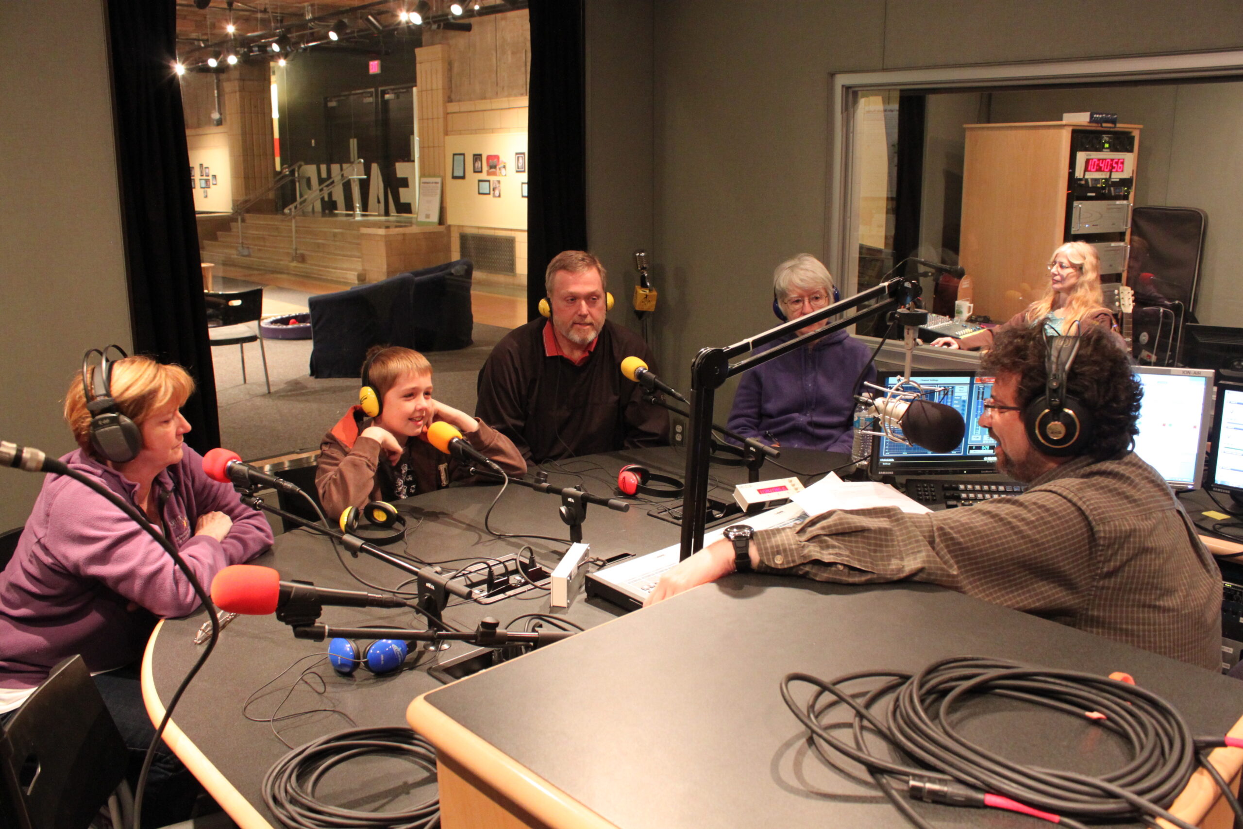 Three adults and a youth wearing headphones speaking into microphones being interviewed by an adult wearing headphones and sitting behind a microphone and an audio mixer in a radio studio