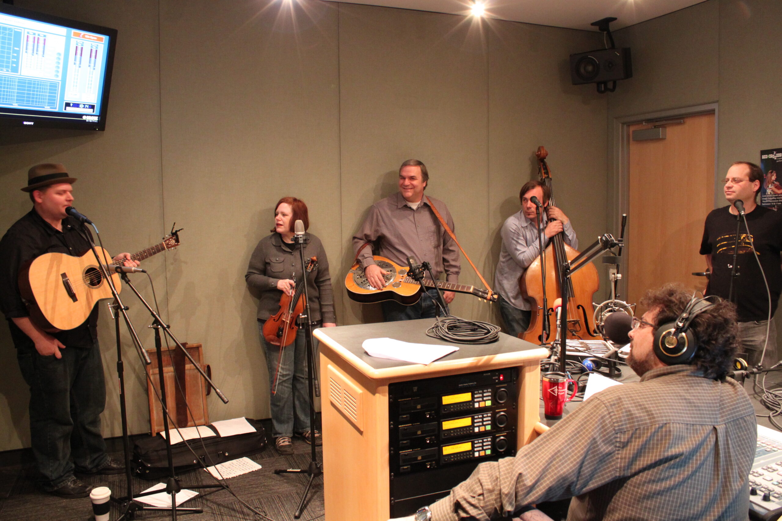 An adult holding an acoustic guitar standing behind a microphone, an adult holding a violin, an adult holding a dobro, an adult holding an upright bass, an adult standing behind a microphone, and an adult wearing headphones sitting behind a microphone in a radio studio