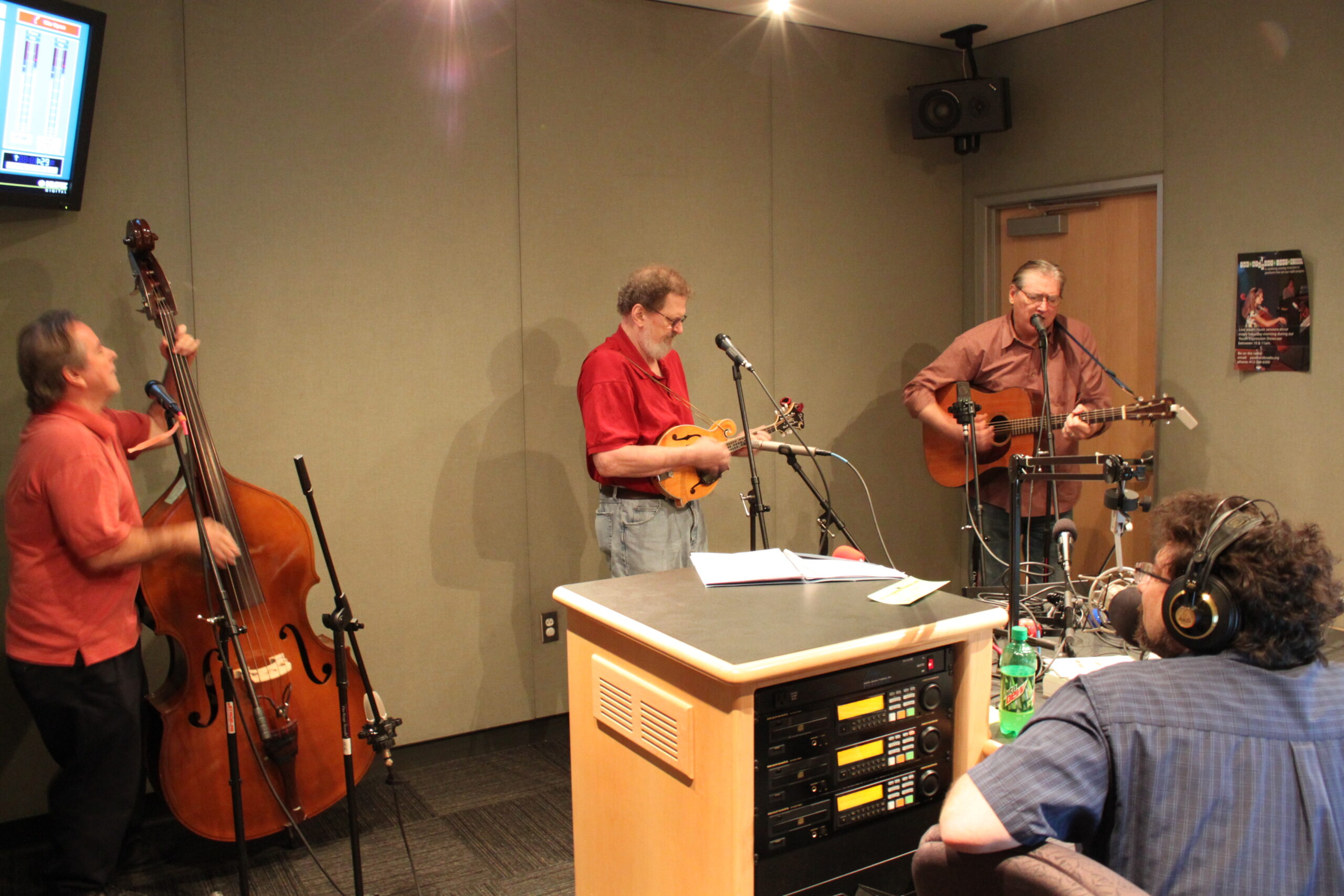 An adult playing upright bass, an adult playing mandolin, and an adult playing acoustic guitar and singing into a microphone as an adult wearing headphones listens in a radio studio