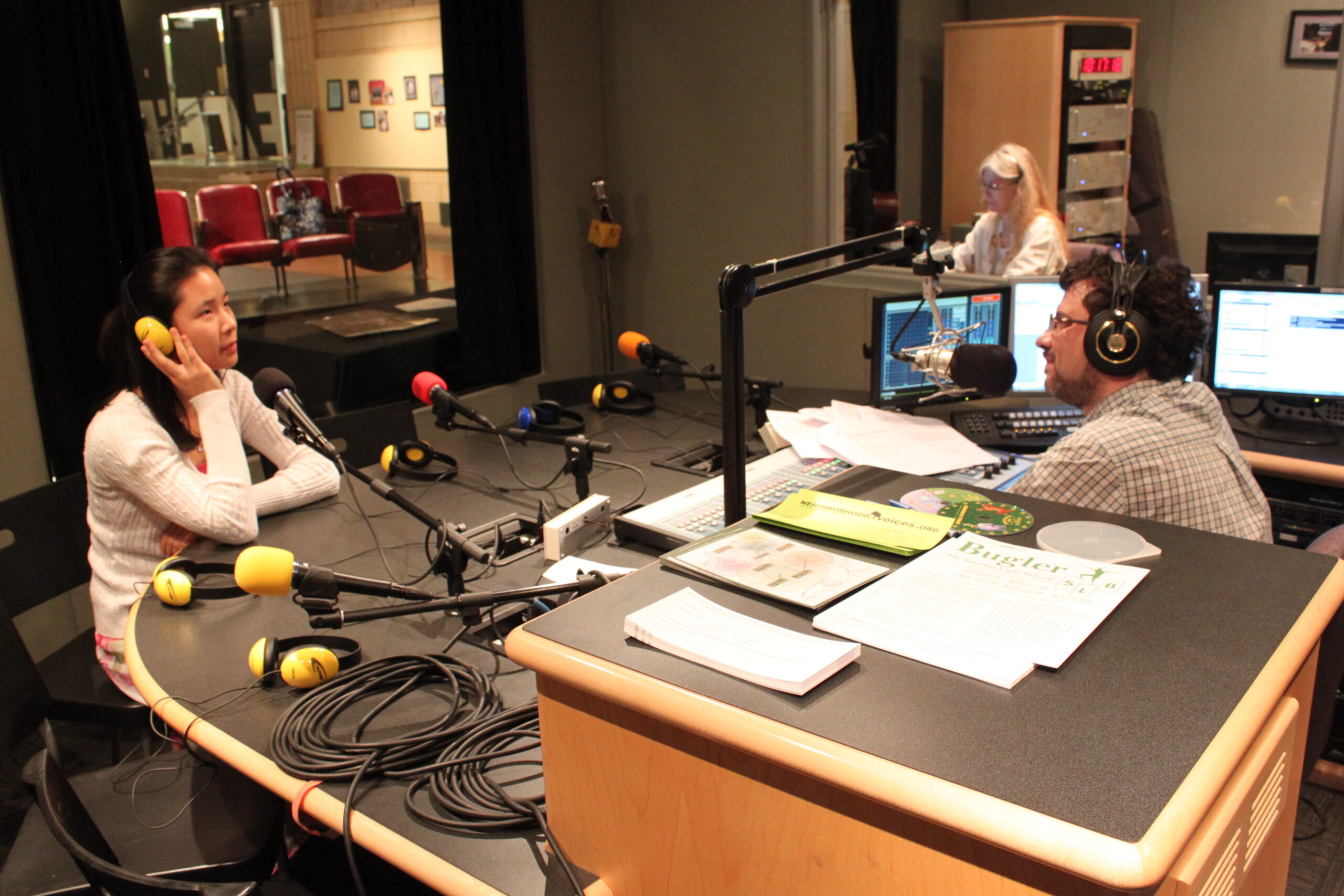 A teen wearing headphones speaking into a microphone being interviewed by an adult wearing headphones sitting behind a microphone and an audio mixer in a radio studio