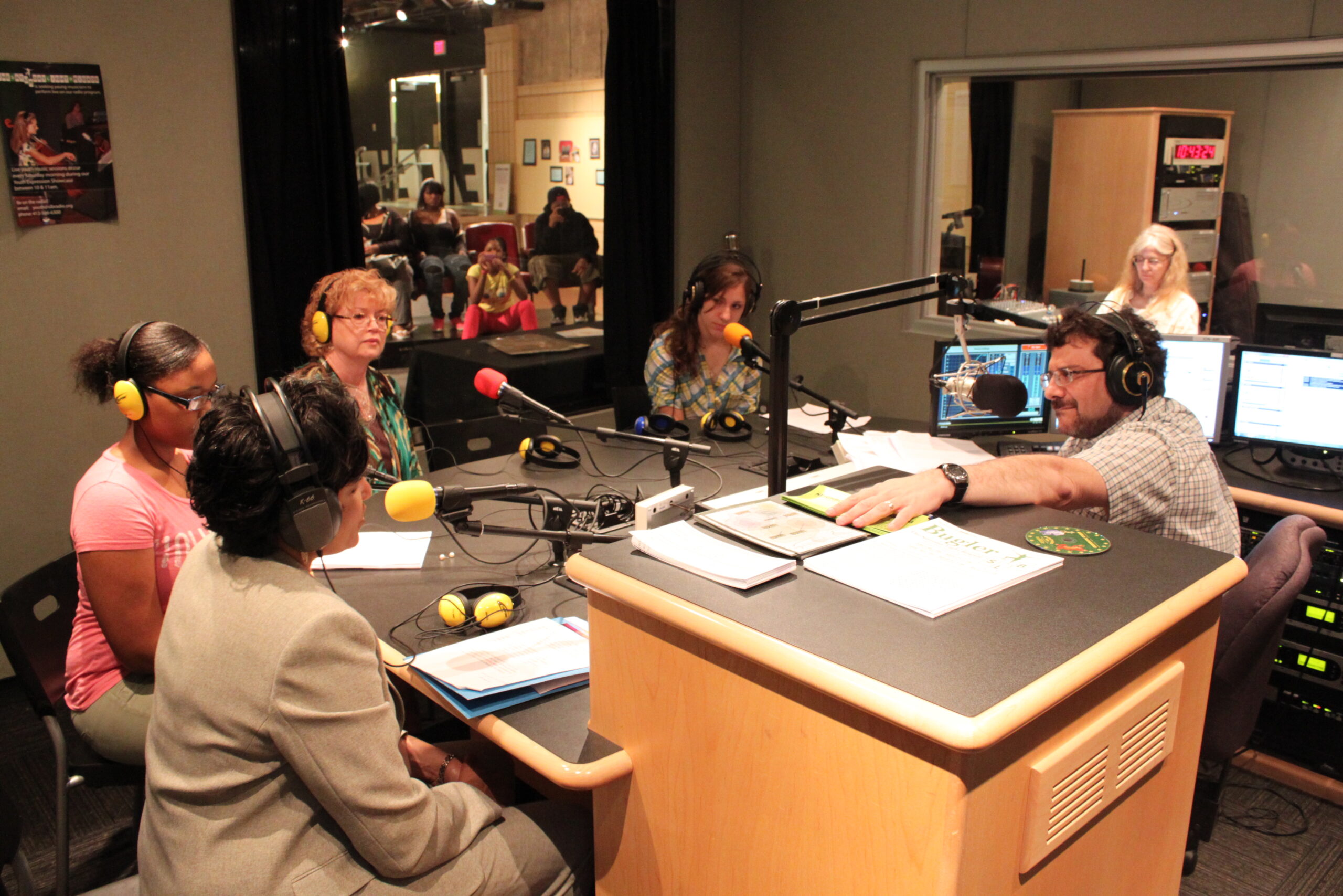 Three teens and one adult wearing headphones speaking into microphones being interviewed by an adult wearing headphones sitting behind a microphone and an audio mixer in a radio studio