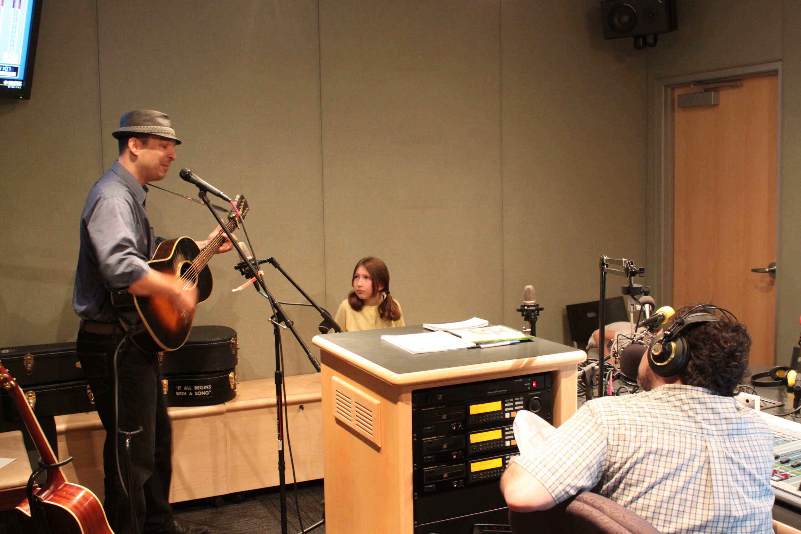 An adult playing an acoustic guitar and singing into a microphone and a youth and an adult listening in a radio studio