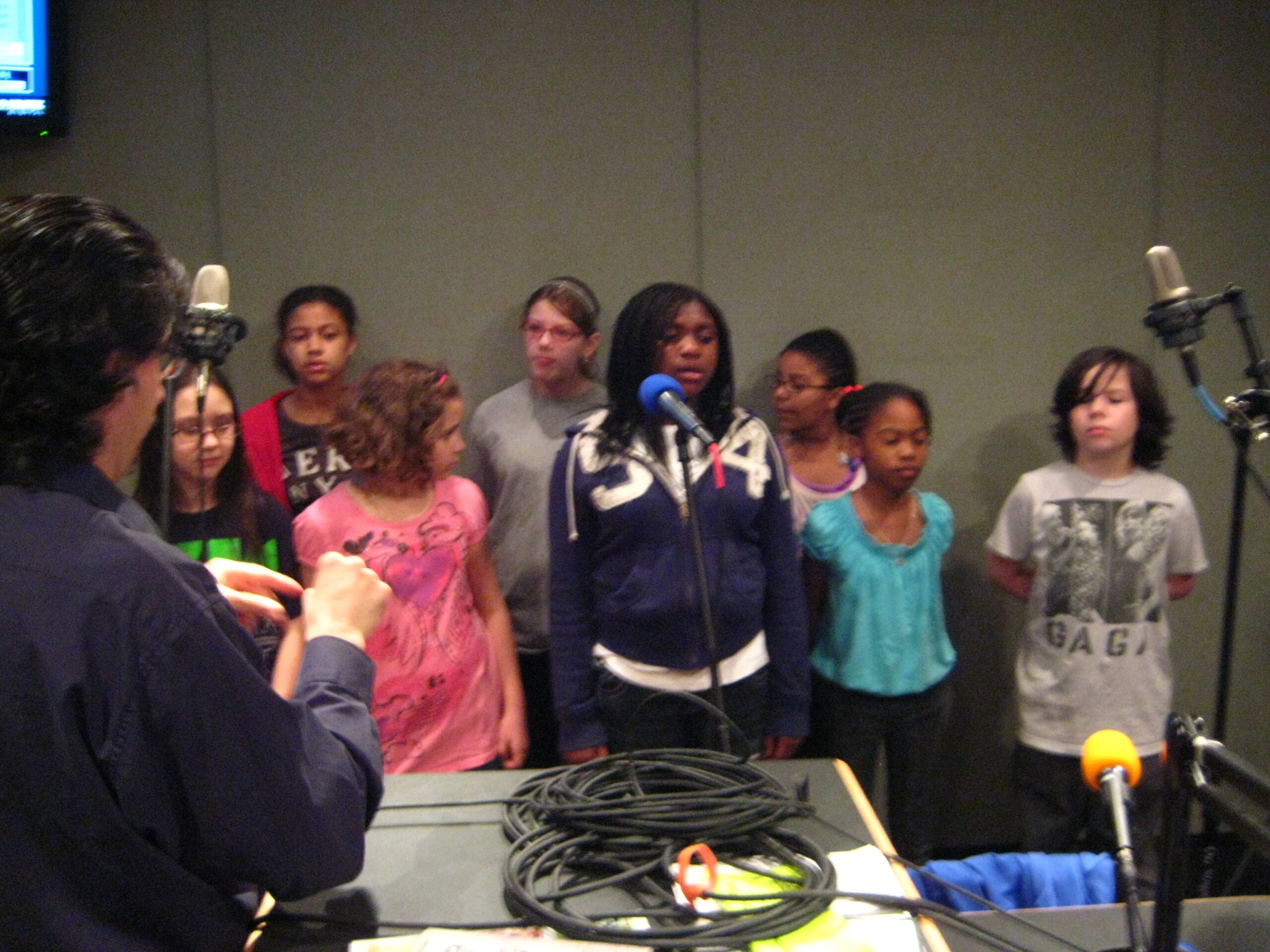 Eight youth singing in a choir being led by an adult in a radio studio